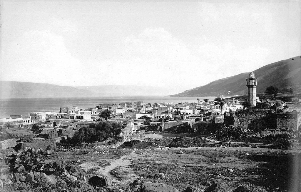 The image depicts a historical view of a coastal town near a body of water, likely a lake or sea, with a prominent lighthouse tower standing tall in the background. The landscape features a mix of stone buildings and ruins, suggesting a blend of architecture that may reflect the area's history. Surrounding the town are hills or mountains, adding depth to the scene. The overall mood is serene and nostalgic, capturing a moment from the past with a monochromatic or sepia tone.