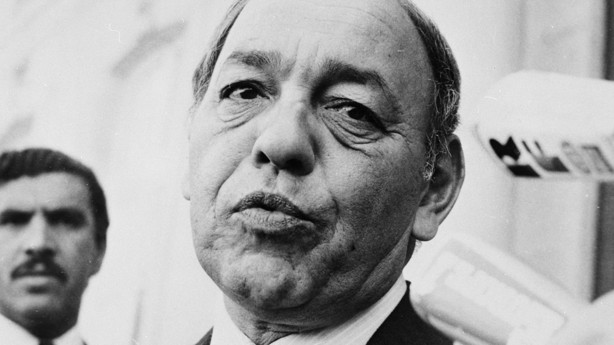 این تصویر یک مرد میانسال را نشان می‌دهد که ظاهری جدی و متمرکز دارد. او لباس رسمی به تن دارد و به دوربین نگاه می‌کند. در پس‌زمینه، شخص دیگری نیز دیده می‌شود که به نظر می‌رسد در حال نظارت یا همراهی با او است. فضای تصویر به طور کلی رسمی و مرتبط با یک موقعیت اجتماعی یا سیاسی به نظر می‌رسد.