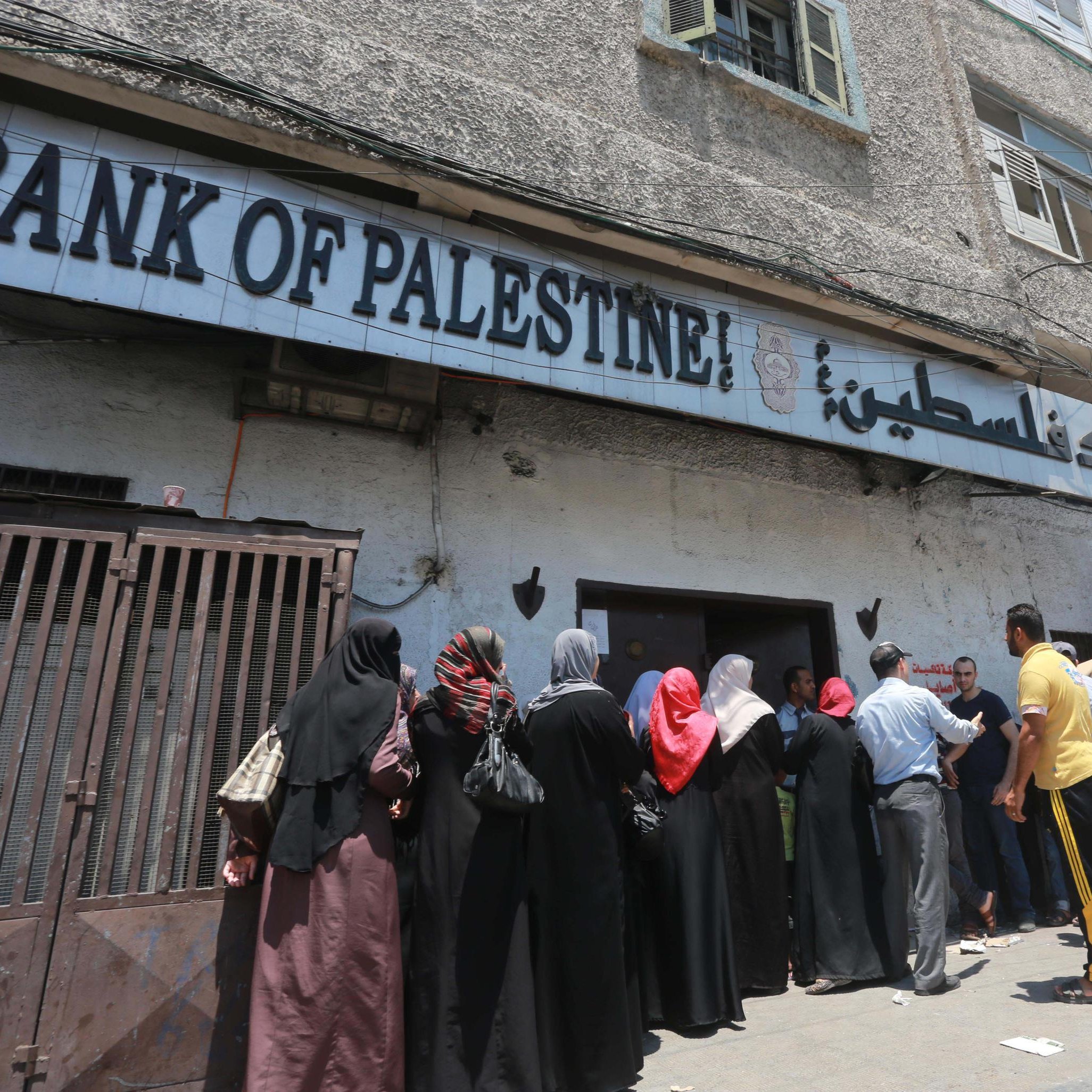 L'image montre une scène extérieure près d'un bâtiment portant l'inscription "Bank of Palestine". On y voit une file de personnes, principalement des femmes portant des vêtements traditionnels, attendant à l'extérieur de la banque. Certains hommes sont également présents, et on aperçoit un groupe d'individus qui se tiennent plus près de l'entrée. En arrière-plan, le bâtiment est ancien et les conditions semblent assez chaotiques, avec des câbles électriques visibles et des fenêtres en état d'usage. Le temps semble ensoleillé, suggérant une ambiance chaude.