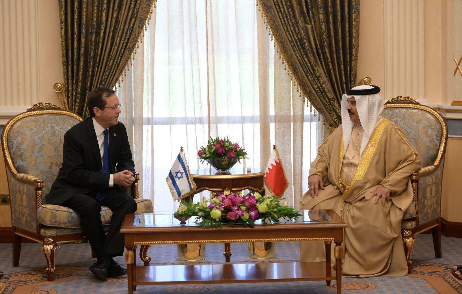 Deux hommes en costume discutent assis dans un salon élégant, drapeaux visibles.