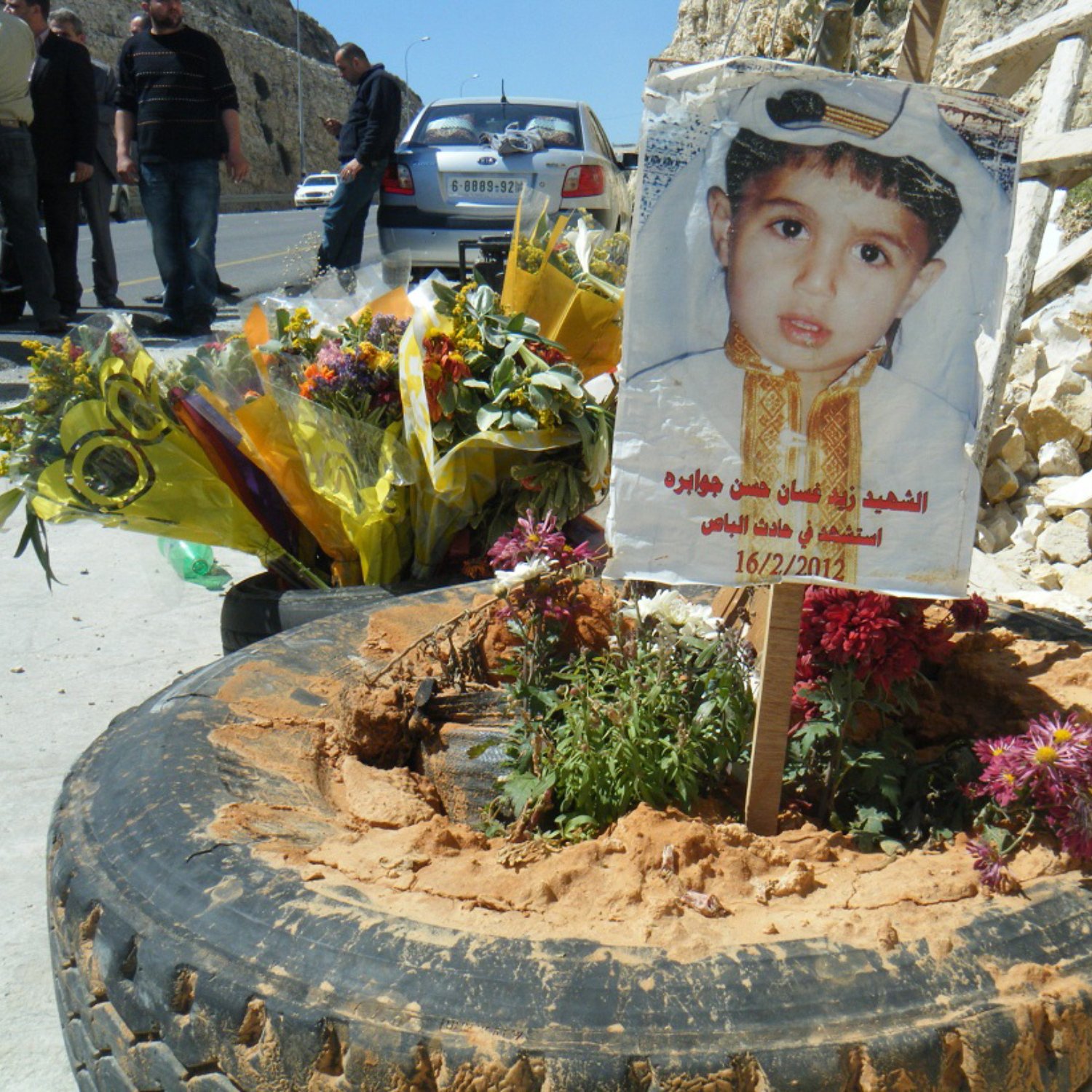 تصویر نشان‌دهنده یک یادبود است که به یاد یک کودک ساخته شده است. در مرکز تصویر، یک دسته گل بزرگ و زیبا قرار دارد و در کنار آن، تابلویی با عکس کودک وجود دارد که لباس خاصی به تن دارد و به تاریخ 16/2/2012 اشاره شده است. زمین اطراف یادبود با شن و گل‌های رنگارنگ تزئین شده و در پس‌زمینه، تعدادی از افراد به نظر می‌رسد که در حال حضور در آن مکان هستند. تصویر احساسات عمیق و یادآوری را منتقل می‌کند.