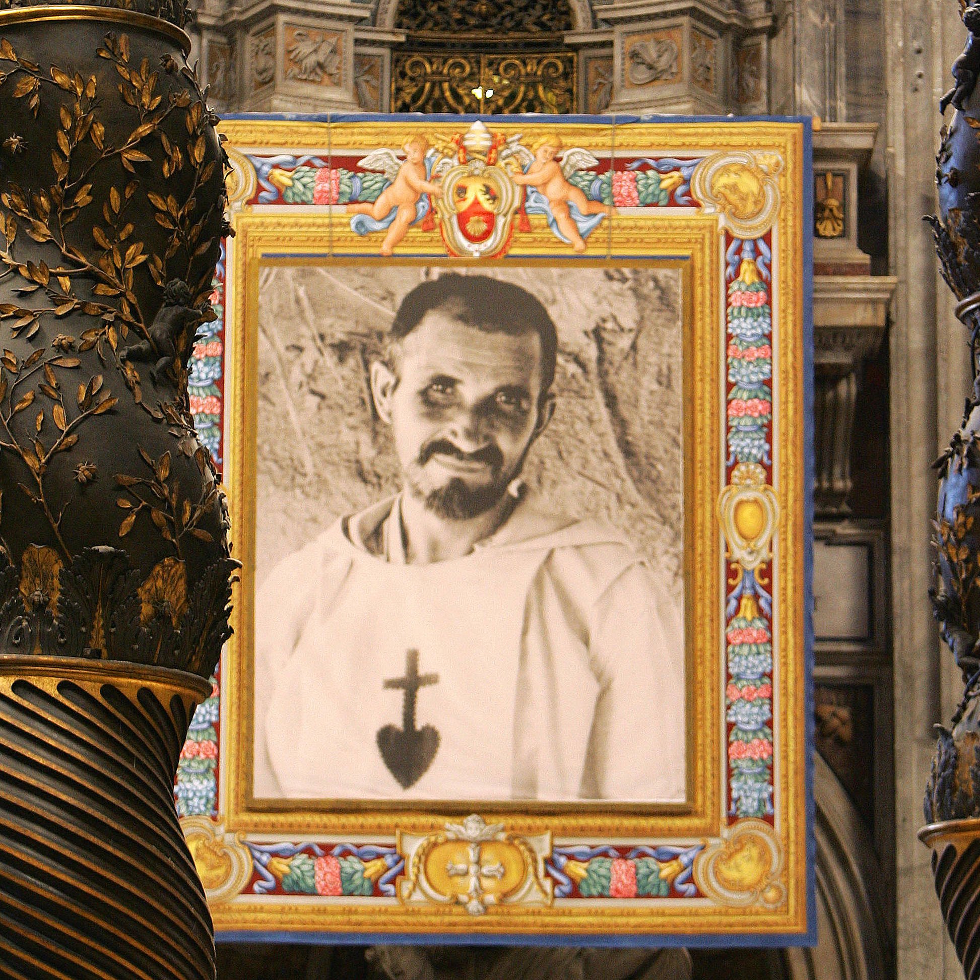 L'image montre un intérieur majestueux, probablement dans une église ou une cathédrale, avec des colonnes ornées de motifs dorés et des détails complexes. Au centre, il y a un grand portrait en noir et blanc d'un homme, qui semble vêtu d'une robe religieuse, encadré de couleurs vives et de décorations. Ce cadre attire l'œil et contraste avec la richesse des ornements de l'architecture environnante. Les murs et les éléments de la pièce évoquent une atmosphère sacrée et historique.