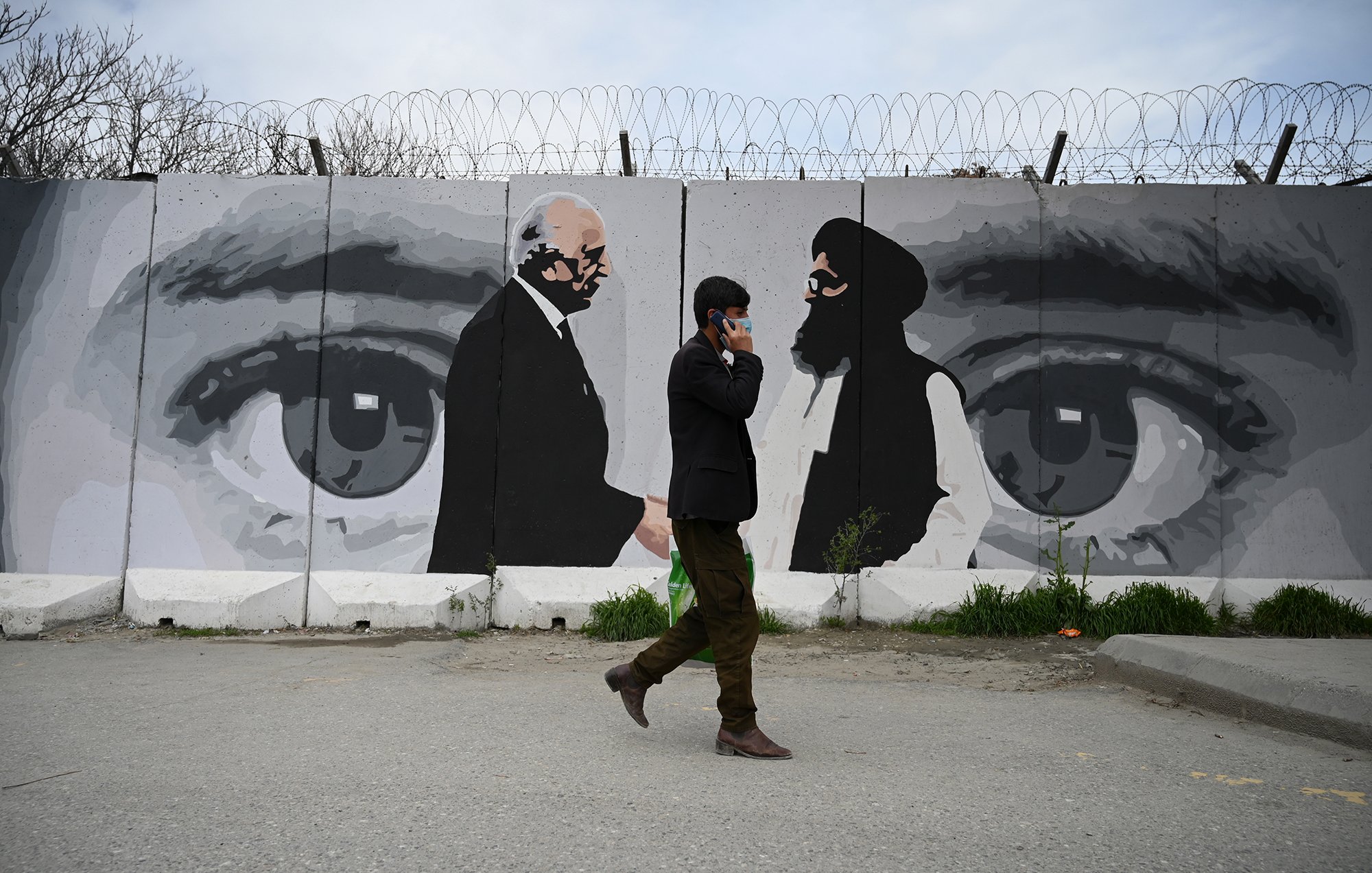 در این تصویر یک مرد در حال راه رفتن در کنار دیواری با یک نقاشی بزرگ است. دیوار دارای تصاویری از دو مرد است که به نظر می‌رسد در حال گفت‌وگو هستند. این نقاشی شامل چشمان بزرگی است که به وضوح بر روی دیوار کشیده شده و احساس عمیقی از توجه و نظارت را منتقل می‌کند. محیط اطراف خیابانی است و مرد به نظر می‌رسد مشغول صحبت کردن با تلفن همراهش باشد. طراحی و رنگ‌ها نسبتاً ملایم و معاصر به نظر می‌رسند.