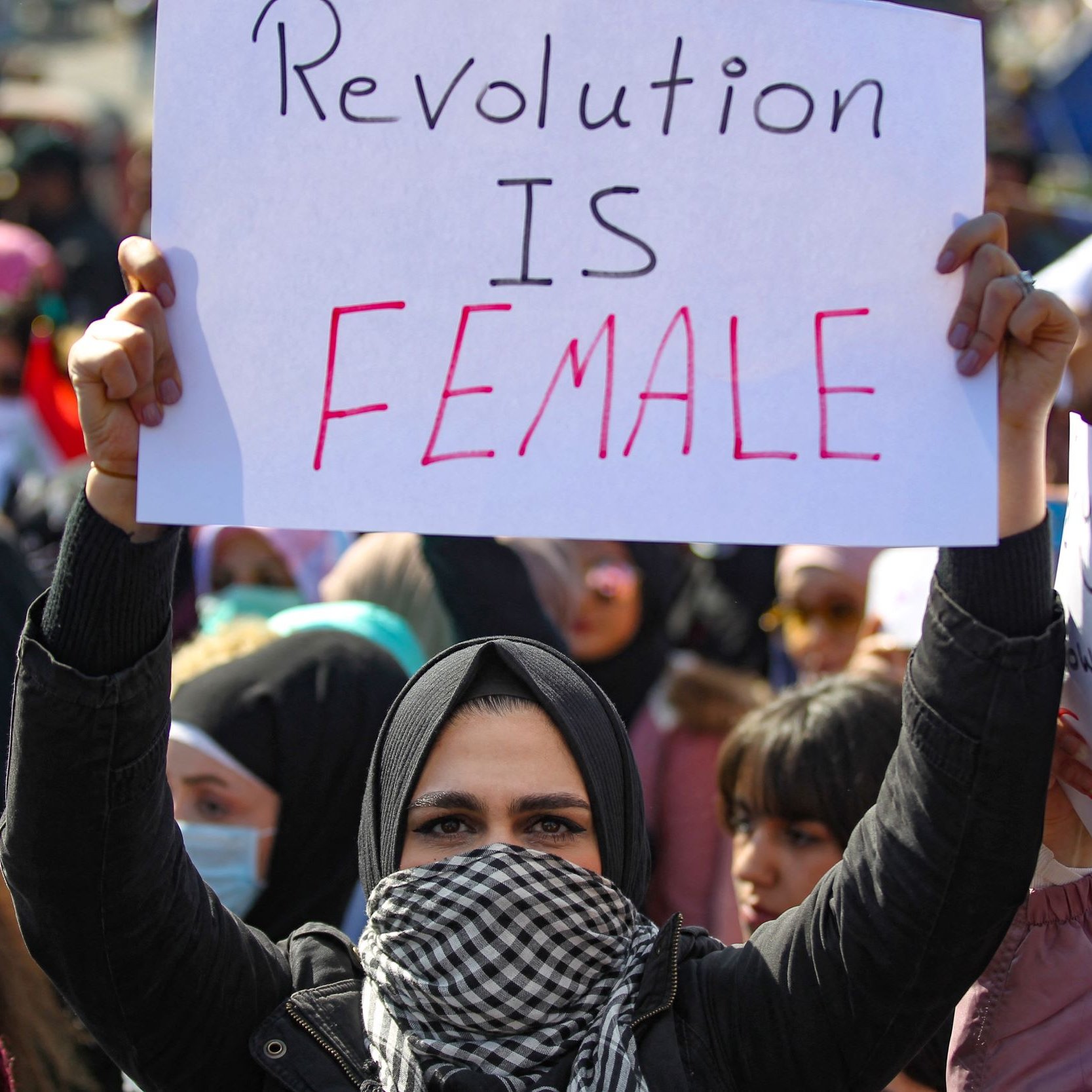 الصورة تظهر مجموعة من المتظاهرين في تجمع حاشد، حيث تحمل امرأة لافتة مكتوب عليها "الثورة أنثوية". يتواجد أفراد من مختلف الأعمار في الخلفية، كما يظهر بعضهم يرتدي الأوشحة والقبعات. الأجواء تعكس حماس المتظاهرين والتزامهم بقضية معينة.