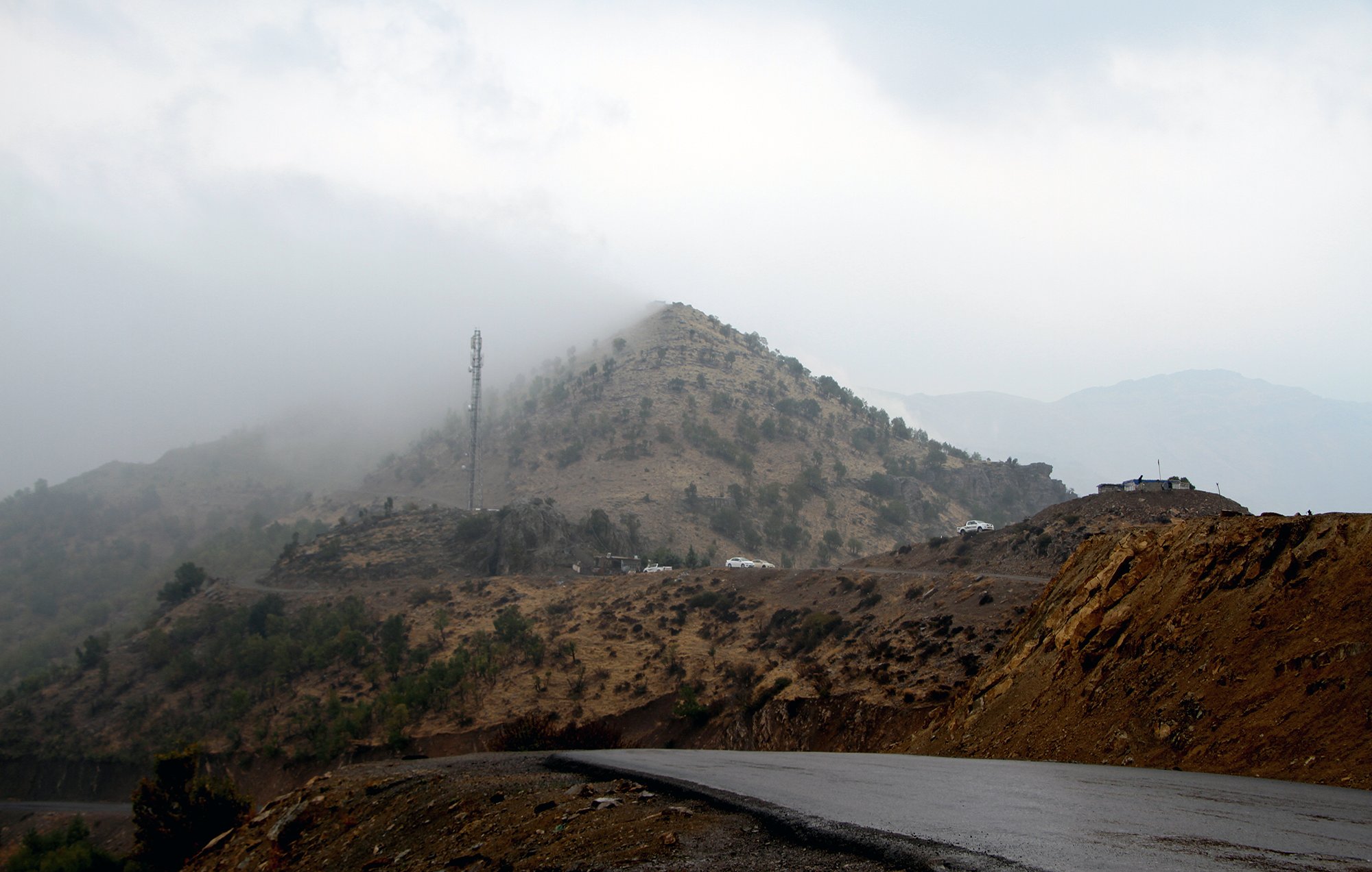 La imagen muestra un paisaje montañoso con una carretera curvada en primer plano. Al fondo, se puede ver una colina cubierta de vegetación escasa y neblina, que le da un aspecto misterioso. En la parte superior de la colina, hay una torre de comunicación. El clima parece nublado y húmedo, creando un ambiente atmosférico y sombrío.
