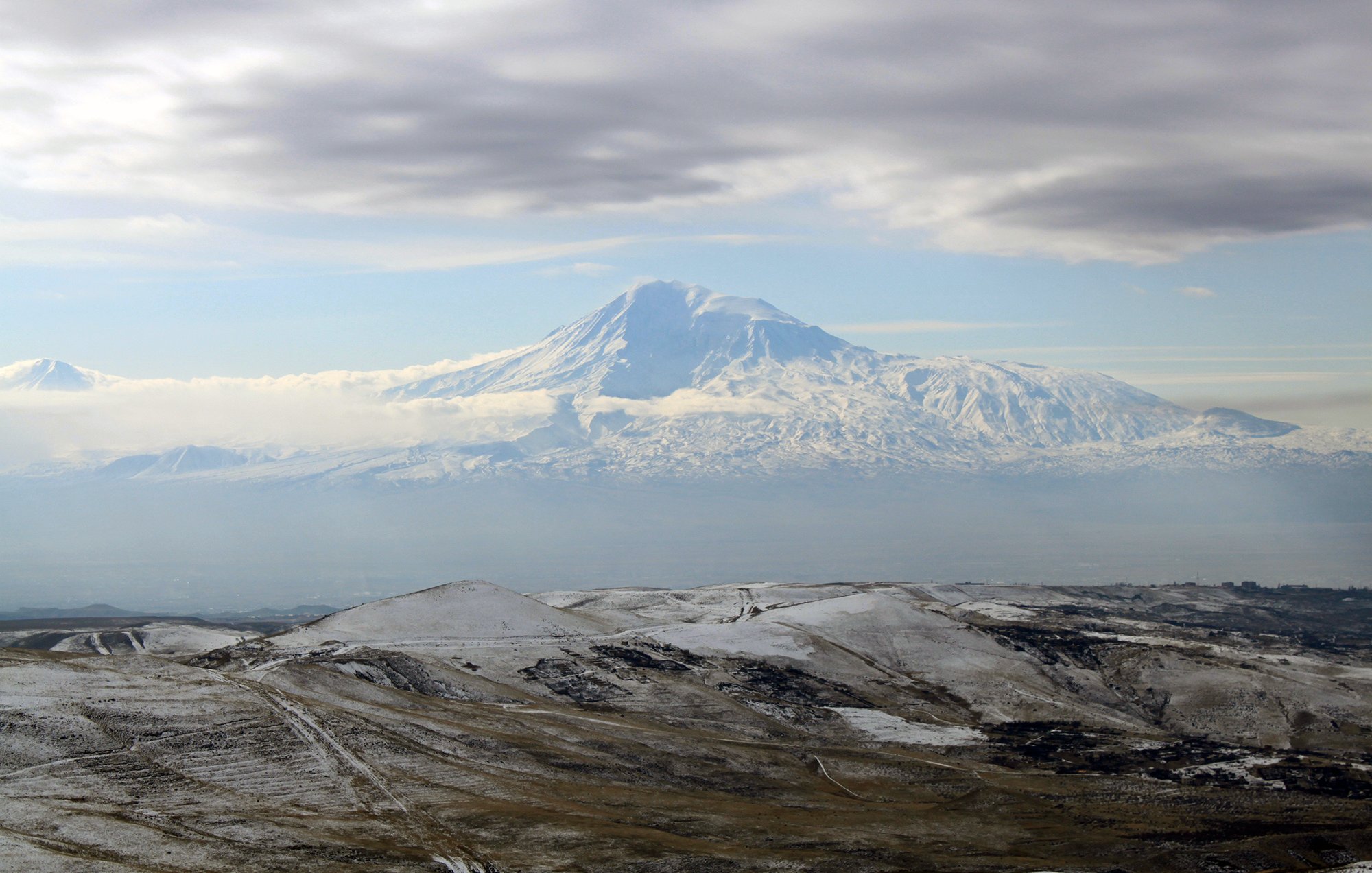 L'image montre un paysage montagneux impressionnant, avec un grand sommet enneigé qui semble être un volcan. Le sommet est entouré de nuages légers, créant une atmosphère mystique. Au premier plan, des collines aux teintes terreuses et parfois enneigées s'étendent, ajoutant à la beauté naturelle de la scène. Le ciel est partiellement couvert, laissant transparaître une lumière douce qui éclaire le paysage.