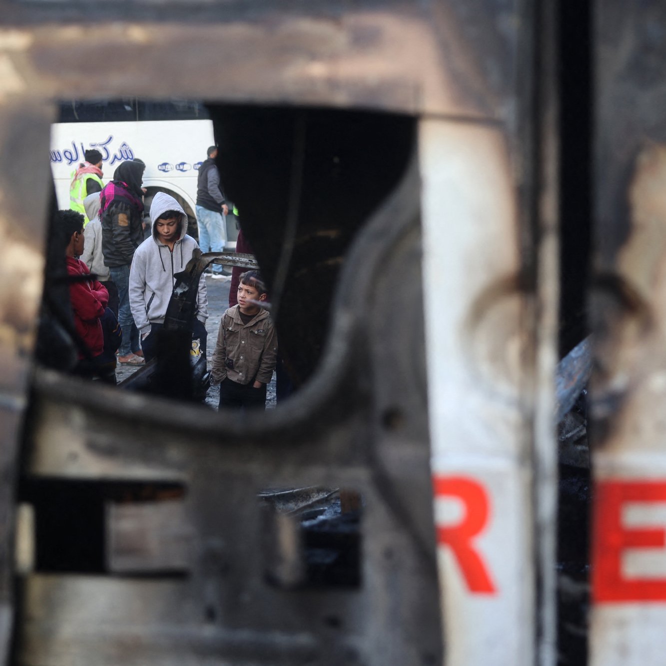 L'image montre un groupe de personnes, y compris des enfants, rassemblées dans un environnement urbain perturbé. On aperçoit une structure endommagée, peut-être un véhicule, qui présente des signes de destruction. Le reflet dans la structure suggère une atmosphère de tension ou de conflit, tandis que les visages des personnes expriment diverses émotions. L'environnement semble chaotique, soulignant une situation difficile.