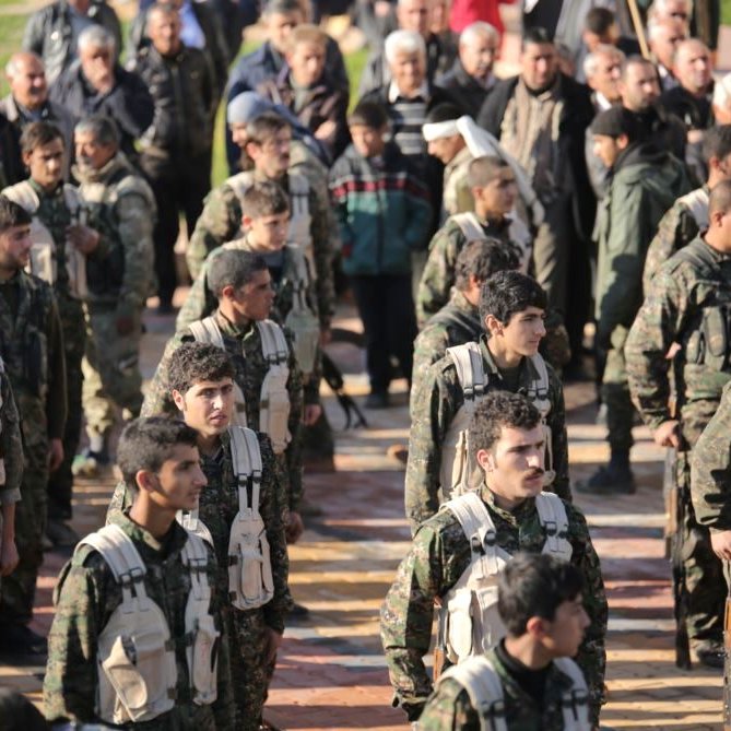L'image montre un groupe de soldats en formation, portant des uniformes militaires et des équipements. Ils sont alignés, en regardant droit devant eux, et semblent être en train de participer à une cérémonie ou à un événement officiel. En arrière-plan, on peut observer une foule de personnes, probablement des civils, qui les regardent. L'atmosphère semble sérieuse et solennelle.