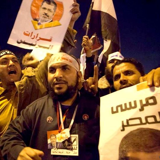 L'image montre un groupe de manifestants engagés dans une démonstration. Ils tiennent des pancartes et des drapeaux, exprimant visiblement leurs opinions politiques. Les visages des participants montrent des émotions intenses, allant de la passion à la détermination. Les slogans et les portraits visibles sur les affiches semblent se rapporter à un leader ou une figure politique, témoignent d'un moment de mobilisation collective. L'ambiance est dynamique, évoquant un fort sentiment de communauté et d'engagement.