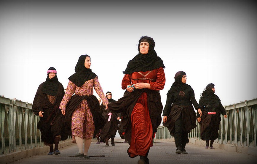 L'image montre un groupe de femmes marchant sur un pont. Elles portent des vêtements traditionnels, principalement des robes longues et des foulards. La scène semble être en extérieur, avec un ciel clair en arrière-plan. Les femmes ont des expressions déterminées et se déplacent ensemble, ce qui dégage une impression d'unité et de force.