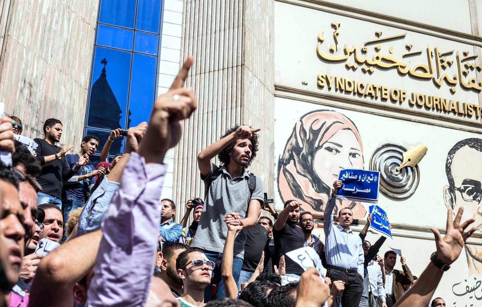 L'image montre une scène de manifestation devant un bâtiment, probablement le Syndicat des journalistes. De nombreuses personnes sont rassemblées, levant les bras en signe de protestation. Certains tiennent des pancartes ou des banderoles, tandis qu'une personne debout sur une foule semble s'adresser aux manifestants. L'arrière-plan présente une grande fresque représentant des figures, ajoutant à l'impact visuel de la scène. L'atmosphère est celle d'un rassemblement engagé et passionné.