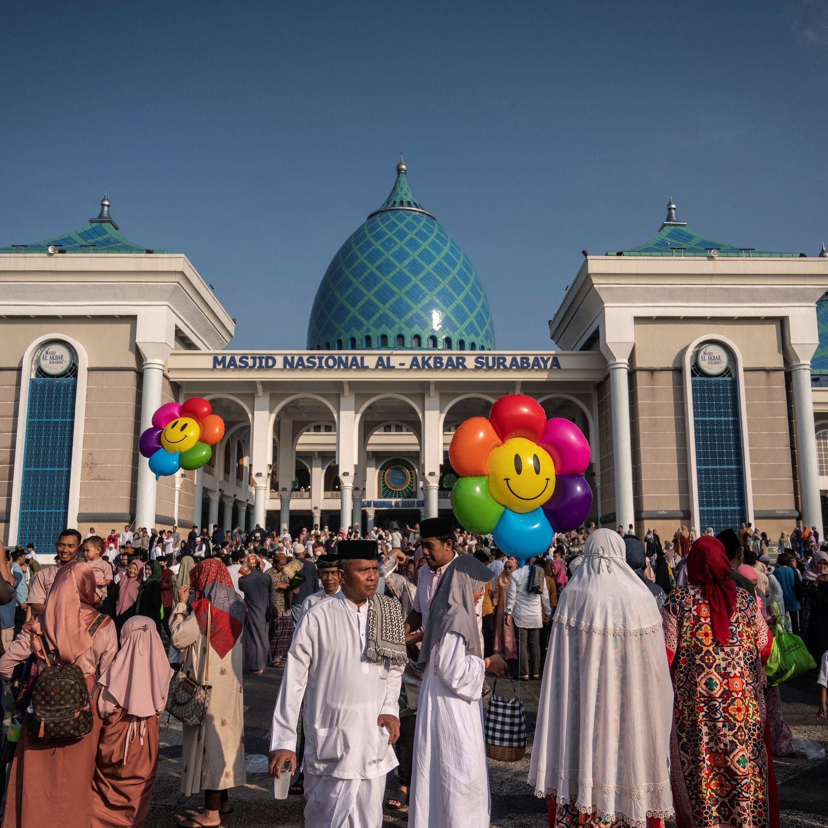 L'image montre un grand rassemblement devant la Masjid Nasional Al-Akbar de Surabaya, en Indonésie. La mosquée est impressionnante, avec des dômes bleu turquoise et une architecture moderne. Beaucoup de personnes sont présentes, certaines portant des vêtements traditionnels, et elles semblent participer à une célébration ou un événement. Des ballons colorés en forme de visages souriants ajoutent une touche festive à la scène. Le ciel est clair, suggérant une belle journée ensoleillée.