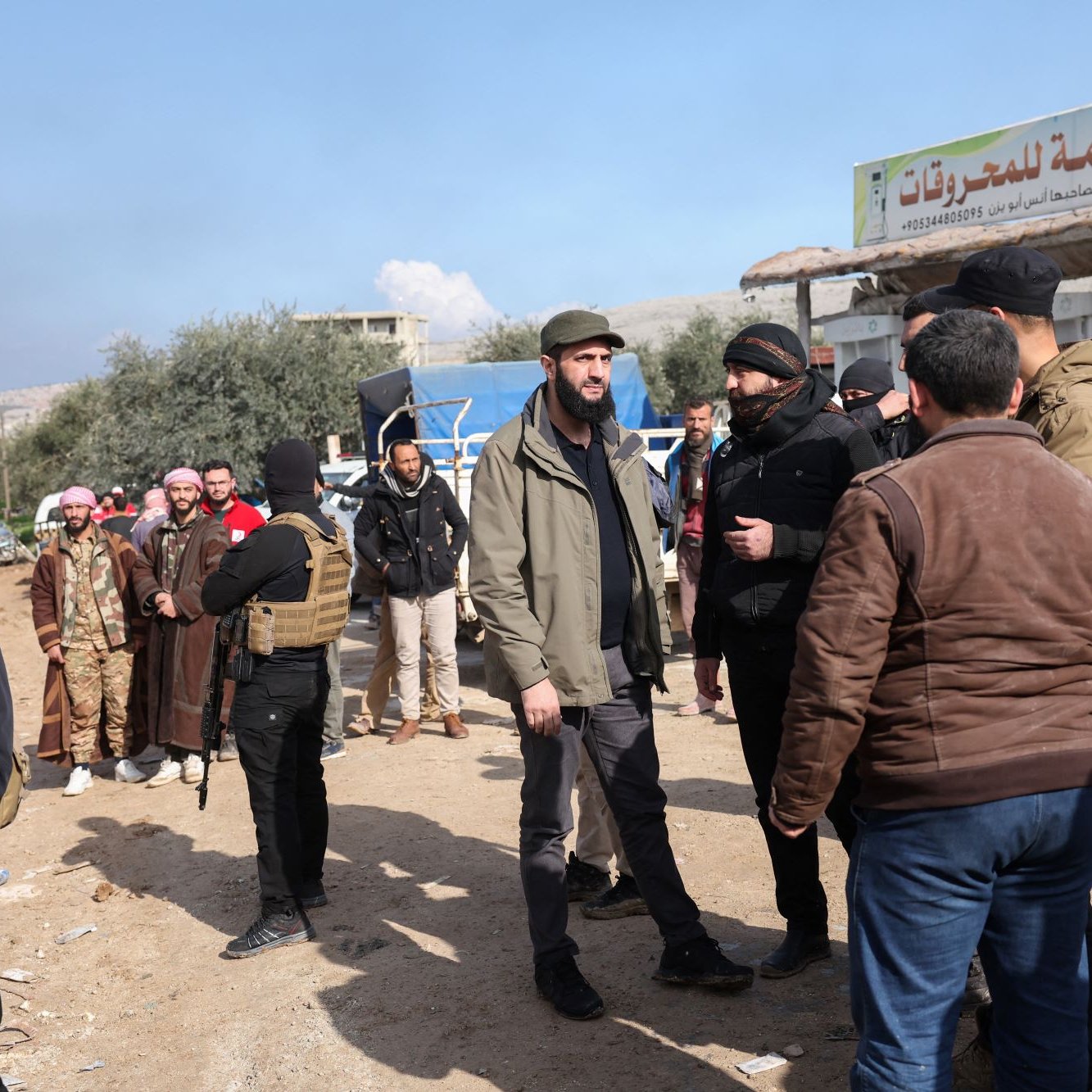 L'image montre un groupe de personnes réunies dans un environnement en plein air, vraisemblablement dans une zone rurale ou semi-urbaine. Certains hommes portent des vêtements traditionnels, tandis que d'autres sont habillés de manière plus moderne. Il y a également des individus en tenue de camouflage avec des gilets tactiques, qui semblent être armés. En arrière-plan, on peut apercevoir un panneau signalétique. Le ciel est partiellement nuageux, ce qui suggère une atmosphère de journée. Les gens interagissent entre eux, ce qui laisse penser qu'il s'agit d'une rencontre ou d'une discussion.
