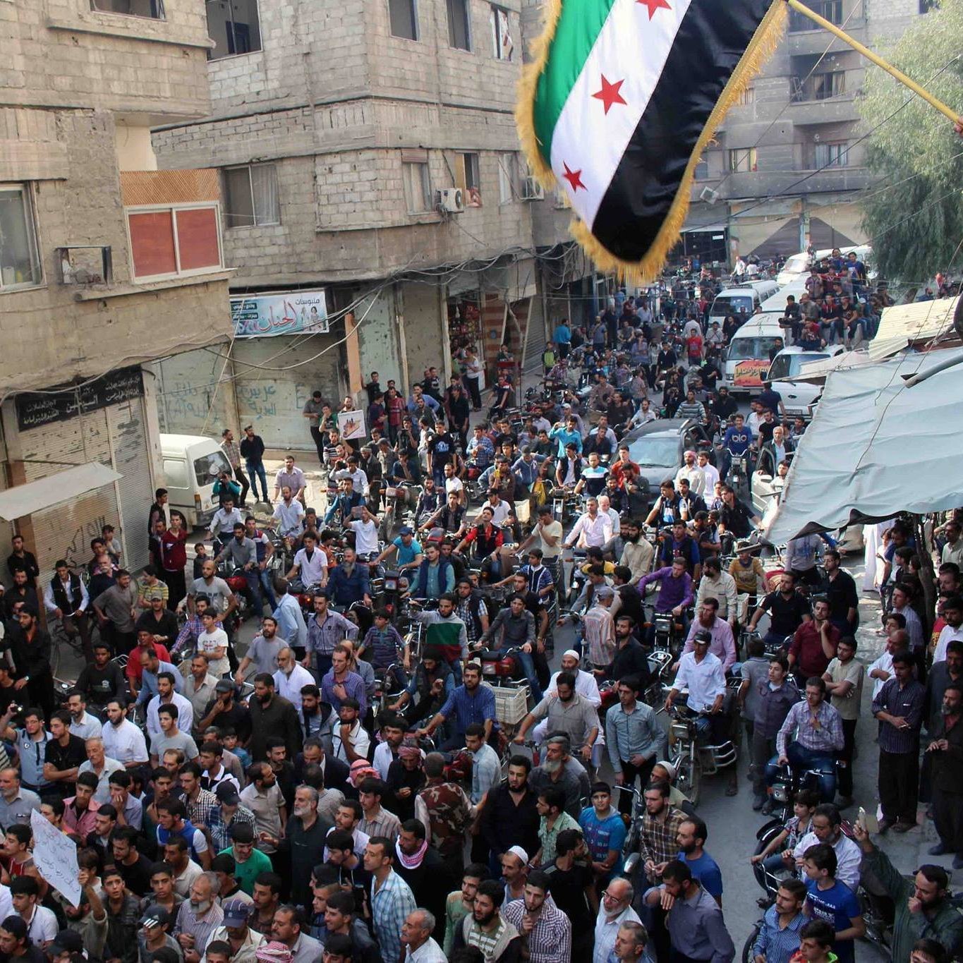 الصورة تظهر حشودًا كبيرة من الناس في شارع مزدحم. يتجمع المحتجون في منطقة حضرية، حيث يمكن رؤية المباني والأسطح والشرفات. علم يرفرف في الأعلى، وهو العلم السوري المعتمد من قبل بعض فصائل المعارضة. يبدو أن هناك جو من الحماس والتجمع، مع وجود بعض السيارات والمركبات في الخلفية.
