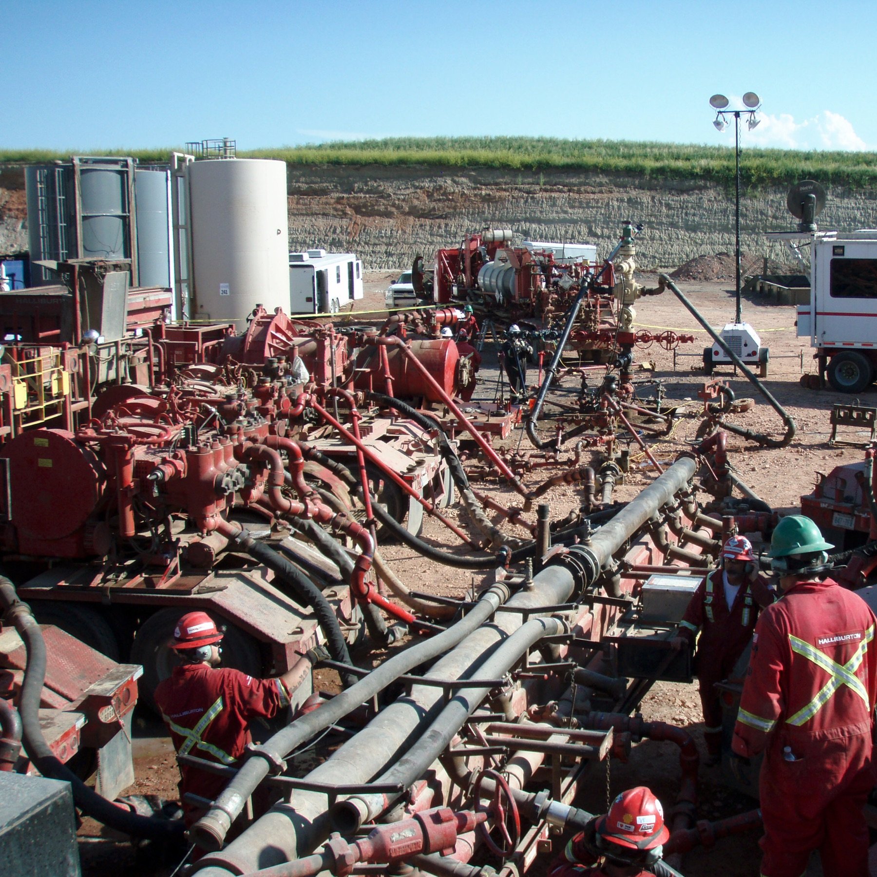 L'image montre un site d'exploitation, probablement lié à l'industrie pétrolière ou gazière. On y voit plusieurs équipements imposants, notamment des pompes et des tuyaux, ainsi que des travailleurs en tenue de sécurité, portant des casques et des vêtements rouges. Le paysage en arrière-plan est rural, avec une colline et des installations industrielles. Le ciel est dégagé, laissant suggérer une journée ensoleillée. L'ensemble de la scène évoque une activité intense et technique, généralement associée à des opérations d'extraction ou de forage.