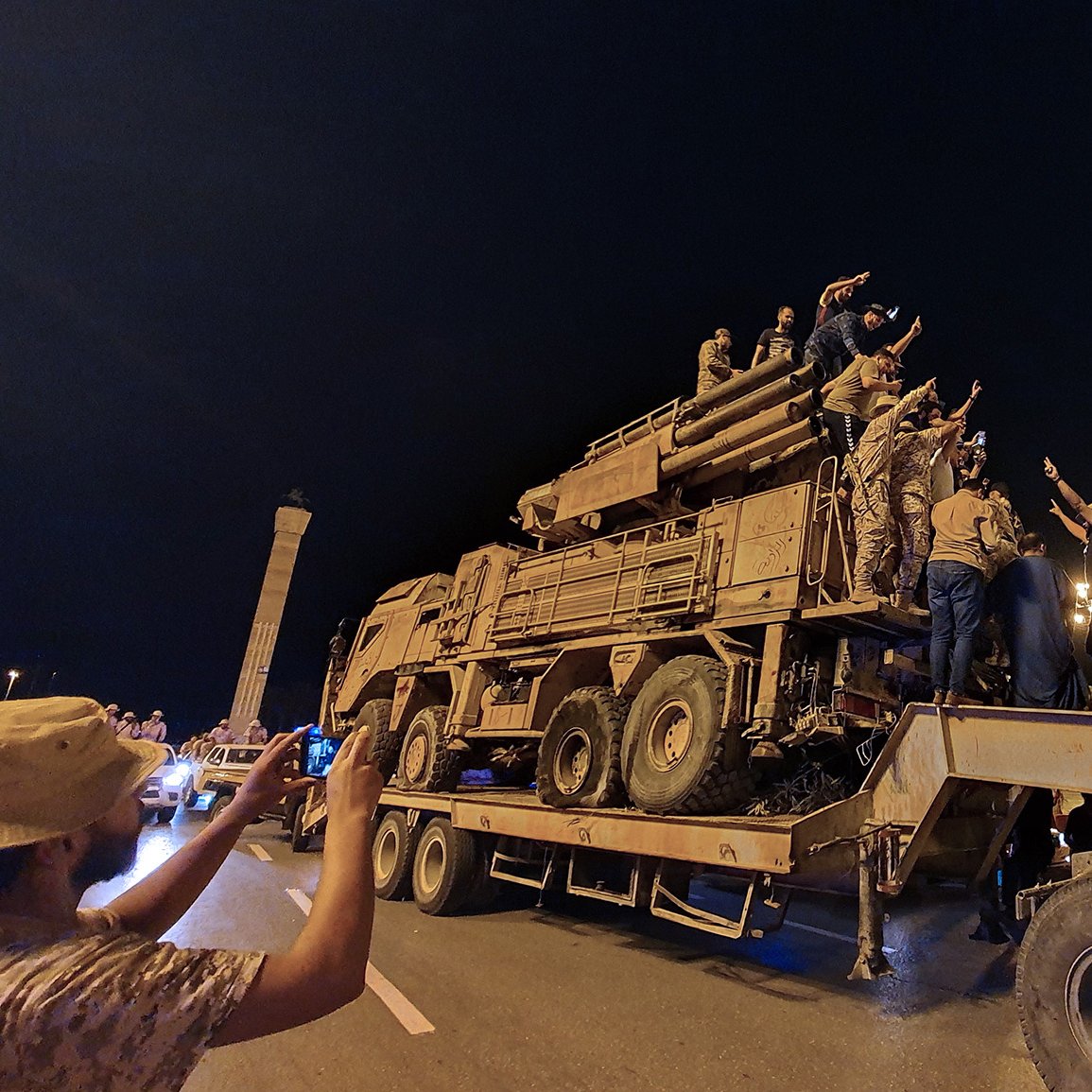 L'immagine mostra un gruppo di persone radunate intorno a un veicolo militare, che sembra essere un camion con un modulo di artiglieria. La scena si svolge di notte, con luci artificiali che illuminano l'ambiente. Alcuni individui sono saliti sopra il veicolo, mentre altri stanno scattando foto con i loro telefoni. L'atmosfera sembra carica di emozione e partecipazione, con gesti di saluto o di celebrazione da parte delle persone sopra il veicolo.