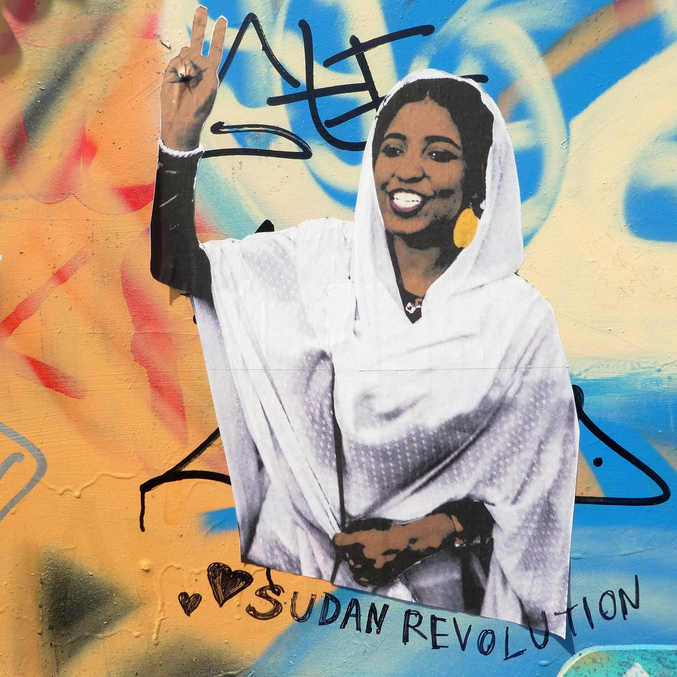 L'image montre une peinture murale colorée sur un mur. Au centre, il y a une représentation d'une femme vêtue d'une robe blanche, levant la main en signe de victoire avec deux doigts. Elle affiche un large sourire, symbolisant la positivité et la résistance. Autour d'elle, il y a des graffitis multicolores, et l'inscription "Sudan Revolution" est bien visible, ce qui indique un lien avec le mouvement révolutionnaire au Soudan. Le tout dégage une ambiance de soutien et de solidarité.