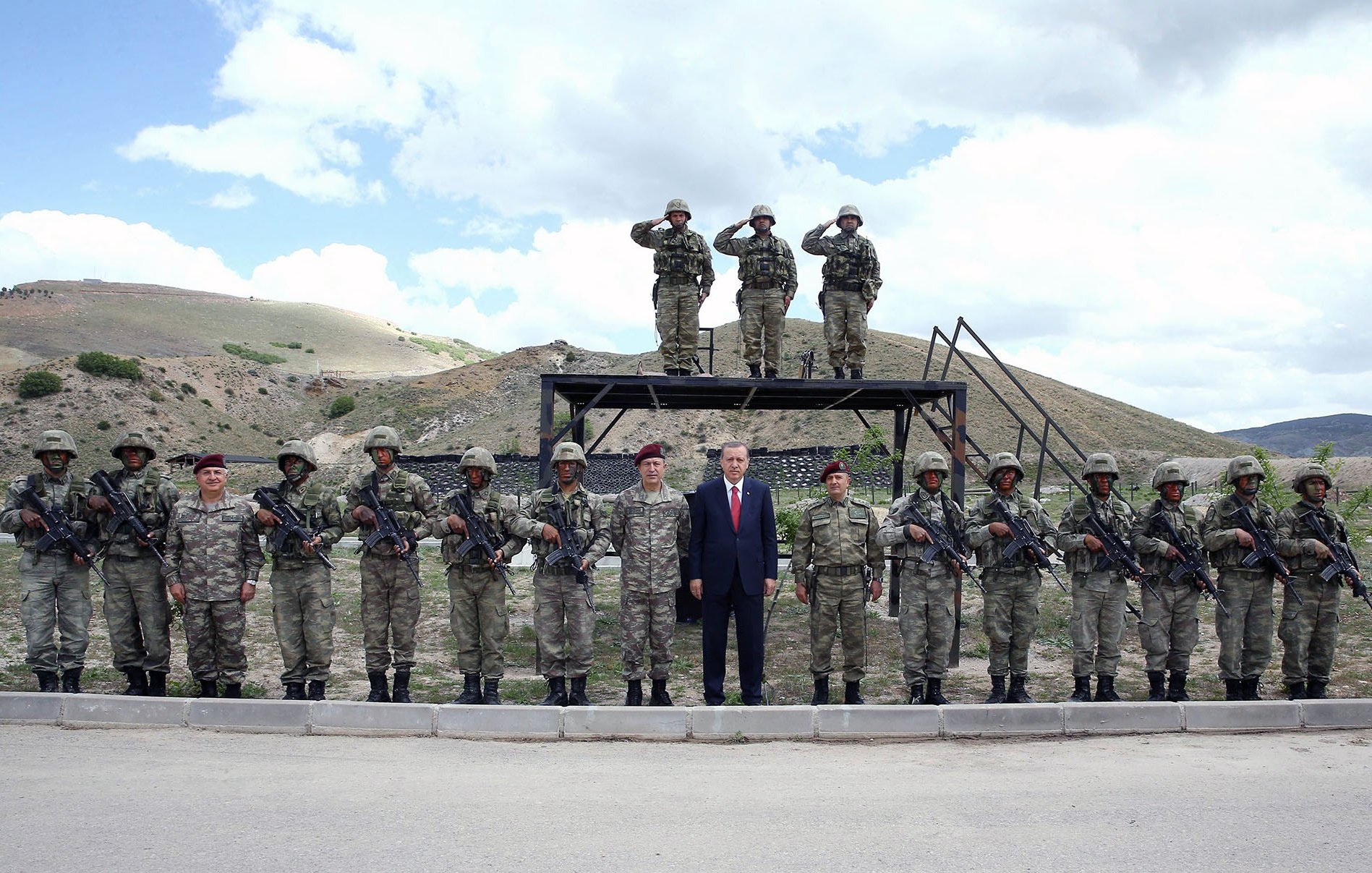 L'image montre un groupe de soldats en uniforme militaire, parfaitement alignés. Au centre, une personne en costume se tient debout parmi eux. À l'arrière-plan, deux soldats saluent en position de hauteur. Les soldats sont armés et entourés d'un paysage montagneux, avec des nuages dans le ciel. L'atmosphère semble formelle et militaire.