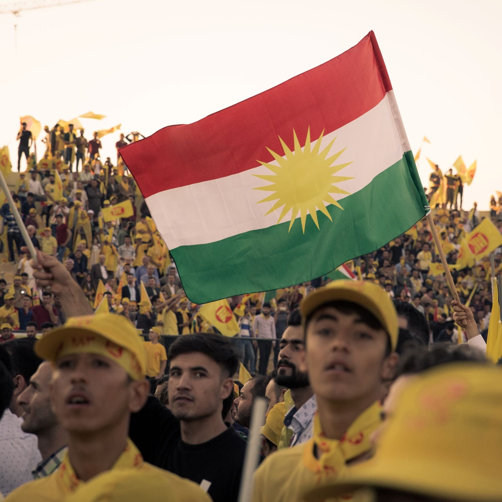 L'immagine mostra un grande raduno di persone che sostengono una causa. In primo piano, un giovane tiene in alto una bandiera con i colori e il simbolo del Kurdistan, che presenta un sole giallo su sfondo rosso e verde. Sullo sfondo, si vedono molte persone, alcune con cappelli o bandiere gialle, che partecipano attivamente all'evento. L'atmosfera è di festa e di unità, con un evidente senso di orgoglio e partecipazione.