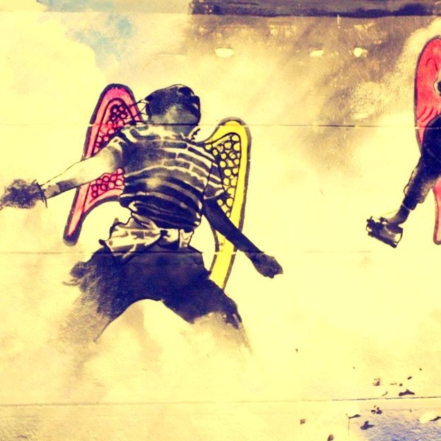 L'image présente deux figures humaines avec des ailes colorées, suggérant une représentation artistique et symbolique. L'une des figures porte un t-shirt rayé, tandis que l'autre est habillée en noir. Les ailes, d'un jaune lumineux pour l'une et d'un rouge pour l'autre, contrastent avec le fond plus sombre et nuageux, créant une atmosphère à la fois dynamique et énigmatique. Cette œuvre semble évoquer des thèmes de liberté et de conflit, donnant une impression de mouvement et d'énergie.