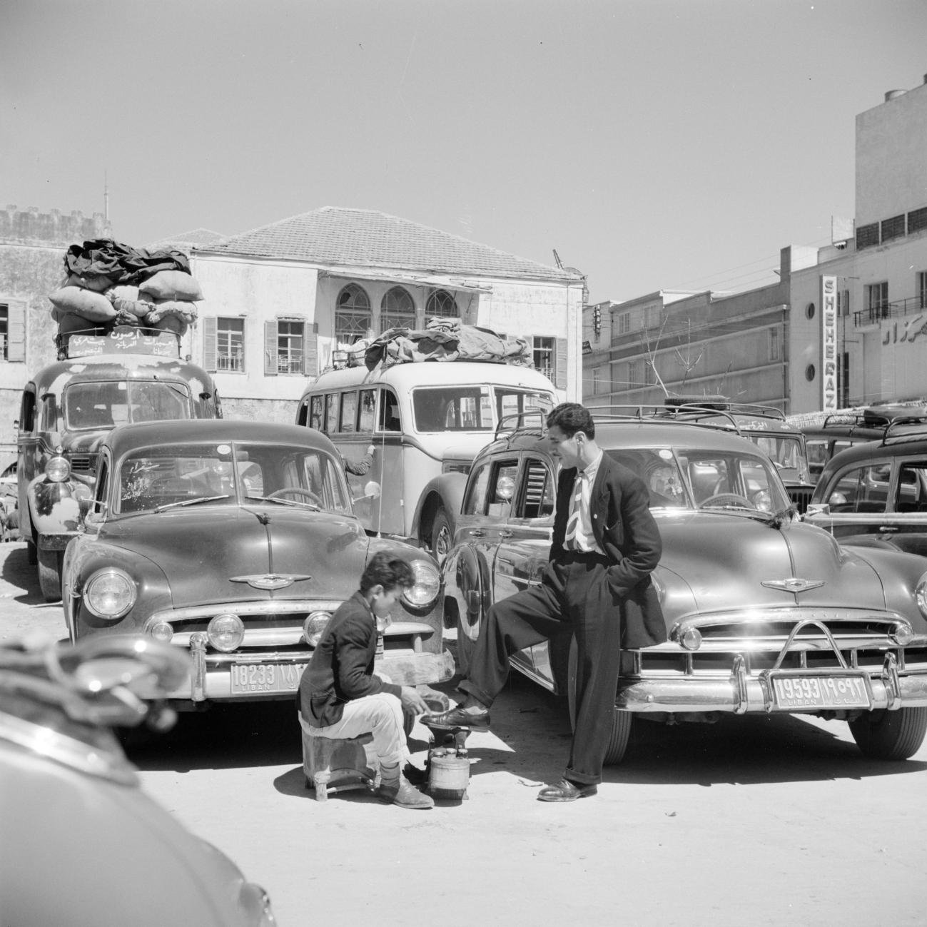 L'image montre une scène de rue animée, probablement dans un contexte urbain d'une époque passée. Au premier plan, un homme en costume se tient debout, l'air décontracté, avec un pied posé sur une petite caisse. Devant lui, un autre homme est accroupi, occupé à préparer quelque chose dans un petit pot ou une casserole. En arrière-plan, on peut voir de nombreuses voitures anciennes, avec des bagages sur leur toit, suggérant peut-être un moment de transition ou de voyage. Le cadre est lumineux et semble capter une ambiance d'activité dynamique, typique des villes dans les décennies passées.