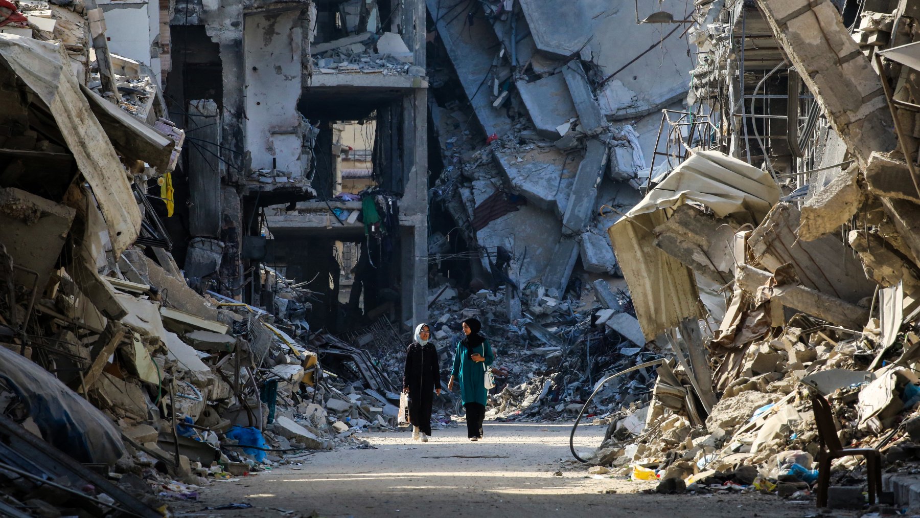 L'immagine mostra due persone che camminano in una strada distrutta da un conflitto, circondate da edifici in rovina. Le strutture sono parzialmente crollate e si vedono macerie sparse ovunque, creando un'atmosfera di desolazione e devastazione. La scena evidenzia la gravità della situazione, con un forte contrasto tra le persone che camminano e l'ambiente circostante, che riflette la difficoltà e la sofferenza di un'area colpita dalla guerra.