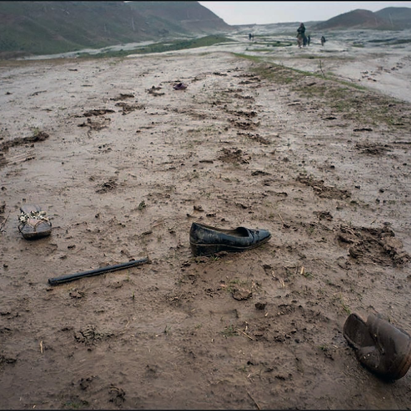 L'image montre un paysage de terre boueuse et humide, probablement après une pluie. On peut voir des chaussures abandonnées, l'une étant une élégante chaussure noire et l'autre, une chaussure brune. Il y a aussi un objet long, semblant être un bâton, sur le sol. En arrière-plan, quelques silhouettes de personnes apparaissent sur le terrain, qui semble montagneux et désolé. L'atmosphère est morose, renforcée par le temps gris et pluvieux.