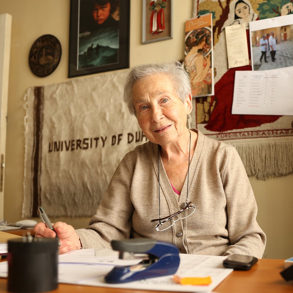 L'image montre une femme âgée assise à un bureau. Elle porte un pull beige et a des lunettes qui reposent sur son nez. Son sourire est chaleureux et accueillant. Le bureau est en désordre avec des papiers éparpillés, un agrafeuse et un verre. En arrière-plan, on aperçoit divers objets accrochés au mur, y compris un calendrier et des photos. On peut aussi distinguer une toile ou un tissu qui mentionne "UNIVERSITY OF DUBAI". L'atmosphère est conviviale et studieuse.