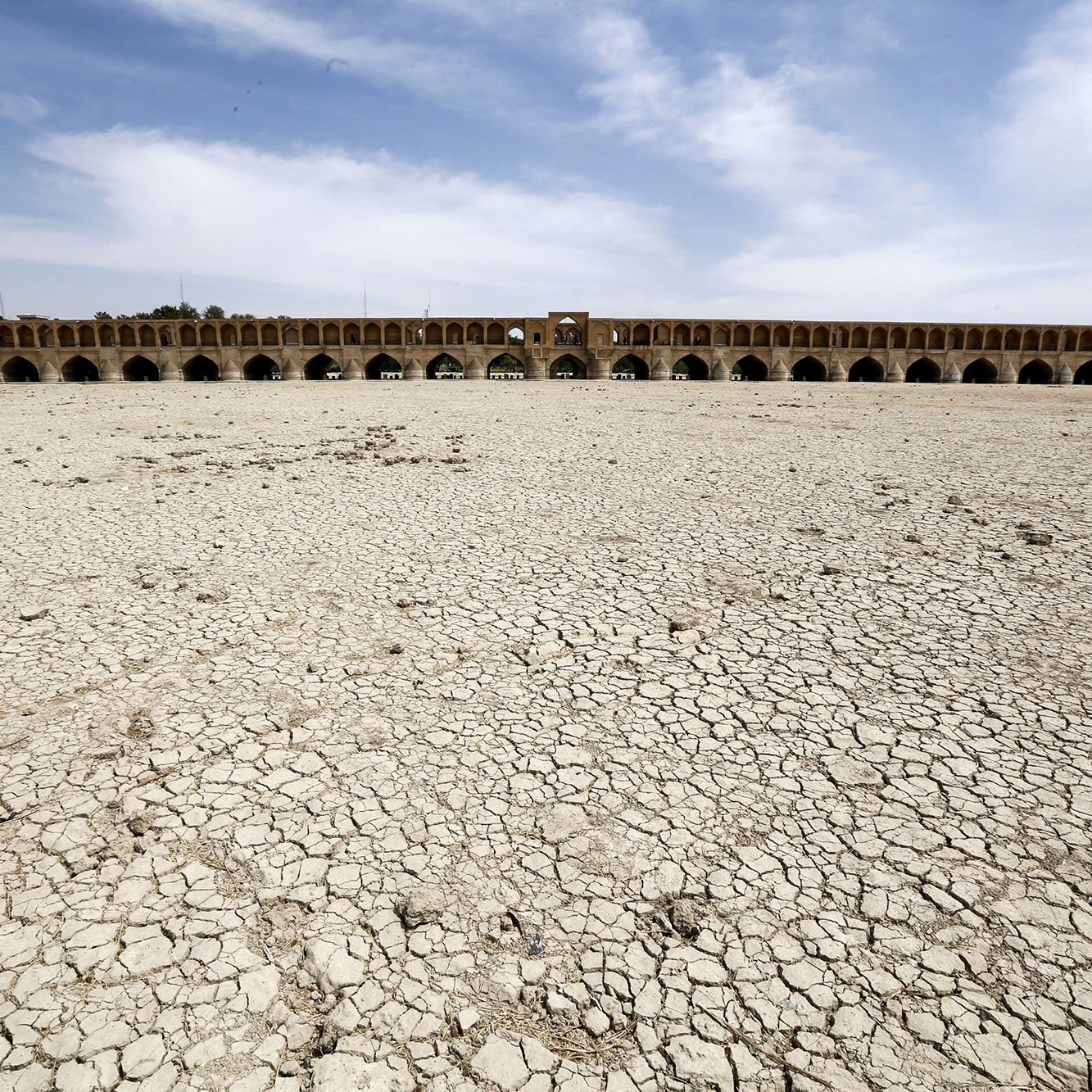 L'image montre un paysage aride avec des sols craquelés, indiquant une sécheresse sévère. À l'arrière-plan, on aperçoit un grand pont historique avec plusieurs arches. Le ciel est dégagé avec quelques nuages, et l'absence d'eau dans la zone suggère des conditions environnementales préoccupantes. Cette scène illustre les effets de la déshydratation sur un espace habituellement aquatique.