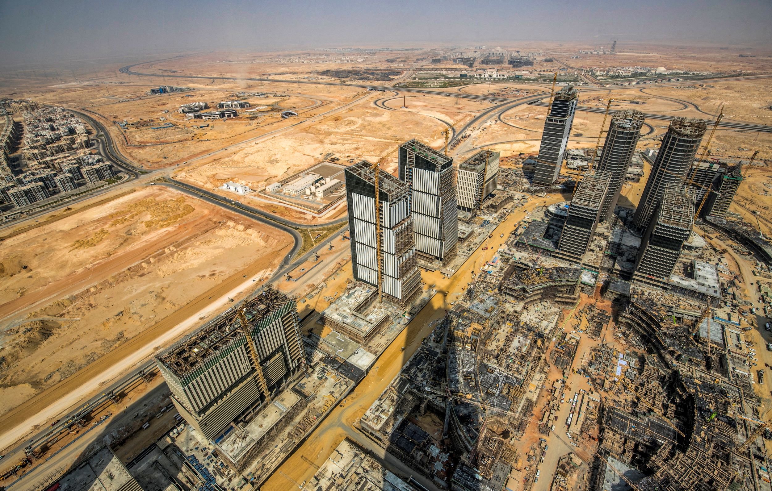 تظهر الصورة منظرًا جويًا لمدينة قيد الإنشاء في منطقة صحراوية. يمكن ملاحظة العديد من الأبراج والمباني الحديثة التي تُشيد، بالإضافة إلى الطرق الواسعة التي تلتف حول المشروع. يحيط بالمكان أرض جرداء، مما يبرز التباين بين البناء الحديث والطبيعة القاحلة. كما تُظهر الصورة التطور العمراني السريع في تلك المنطقة.