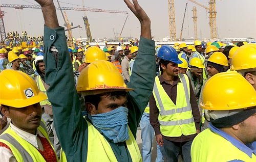 L'image montre un grand groupe de travailleurs, majoritairement en train de lever les bras. Tous portent des casques de sécurité jaunes et des gilets réfléchissants. L'environnement semble être un site de construction, avec plusieurs grues et équipements visibles en arrière-plan. L'atmosphère paraît dynamique et l'affluence des travailleurs indique probablement une activité importante sur le chantier.
