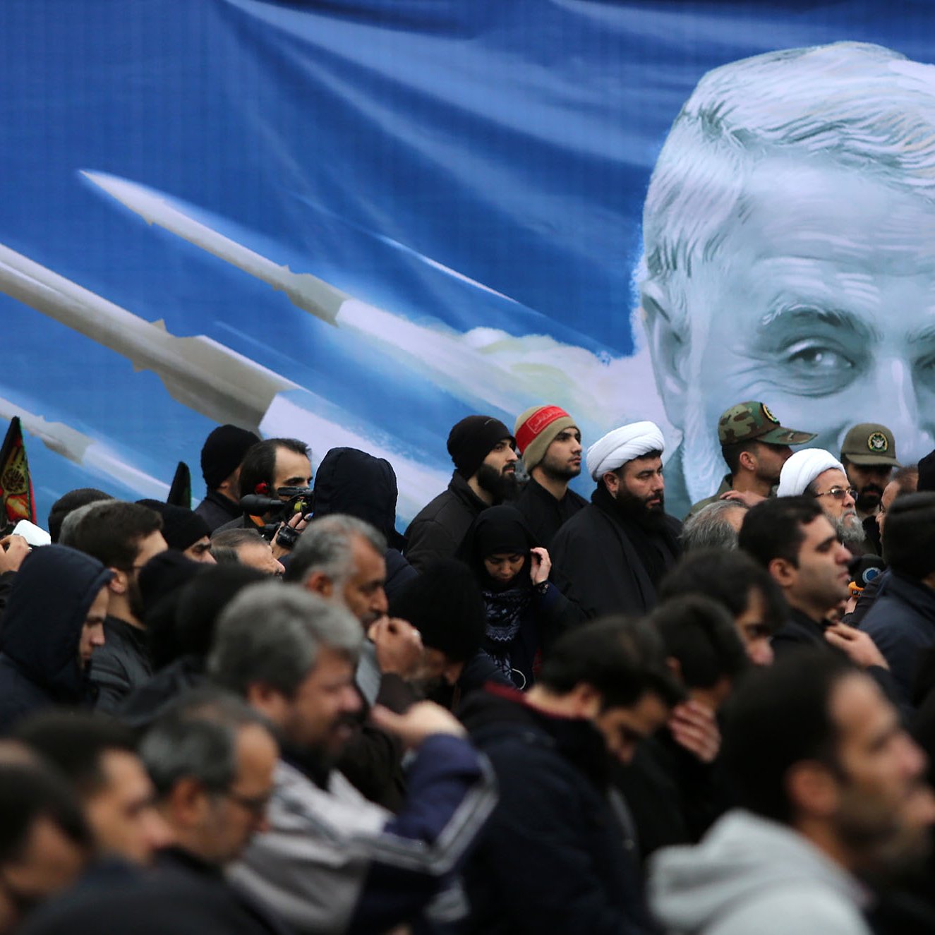 L'image montre une foule de personnes rassemblées, vraisemblablement lors d'un événement public. Les individus portent des vêtements sombres et certains semblent tenir des drapeaux ou des bannières. En arrière-plan, il y a une grande illustration d'un visage avec des images de missiles, ce qui pourrait indiquer un contexte politique ou militaire. L'ambiance générale semble sérieuse et solennelle.