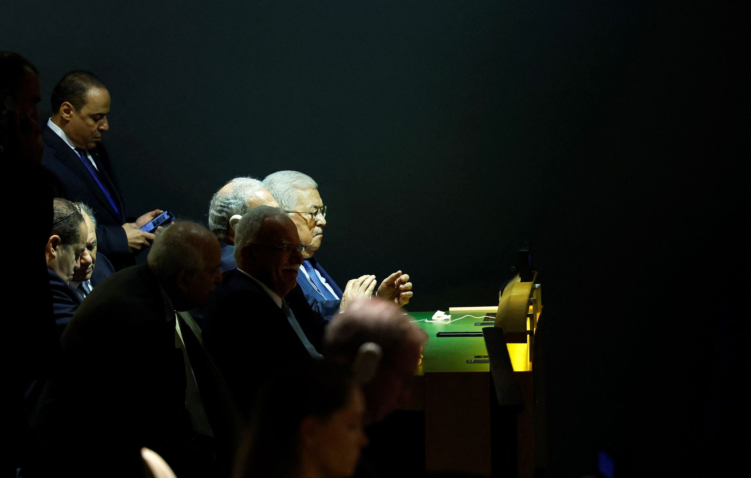 L'immagine mostra un gruppo di uomini seduti a un tavolo, apparentemente impegnati in una discussione o in un evento ufficiale. Alcuni di loro sembrano concentrati, mentre uno di loro sta guardando il cellulare. L'illuminazione è tenue, con un contrasto tra le zone illuminate e quelle in ombra, conferendo un'atmosfera seria e formale all'ambiente. Non ci sono elementi di identificazione chiara per i volti dei partecipanti.