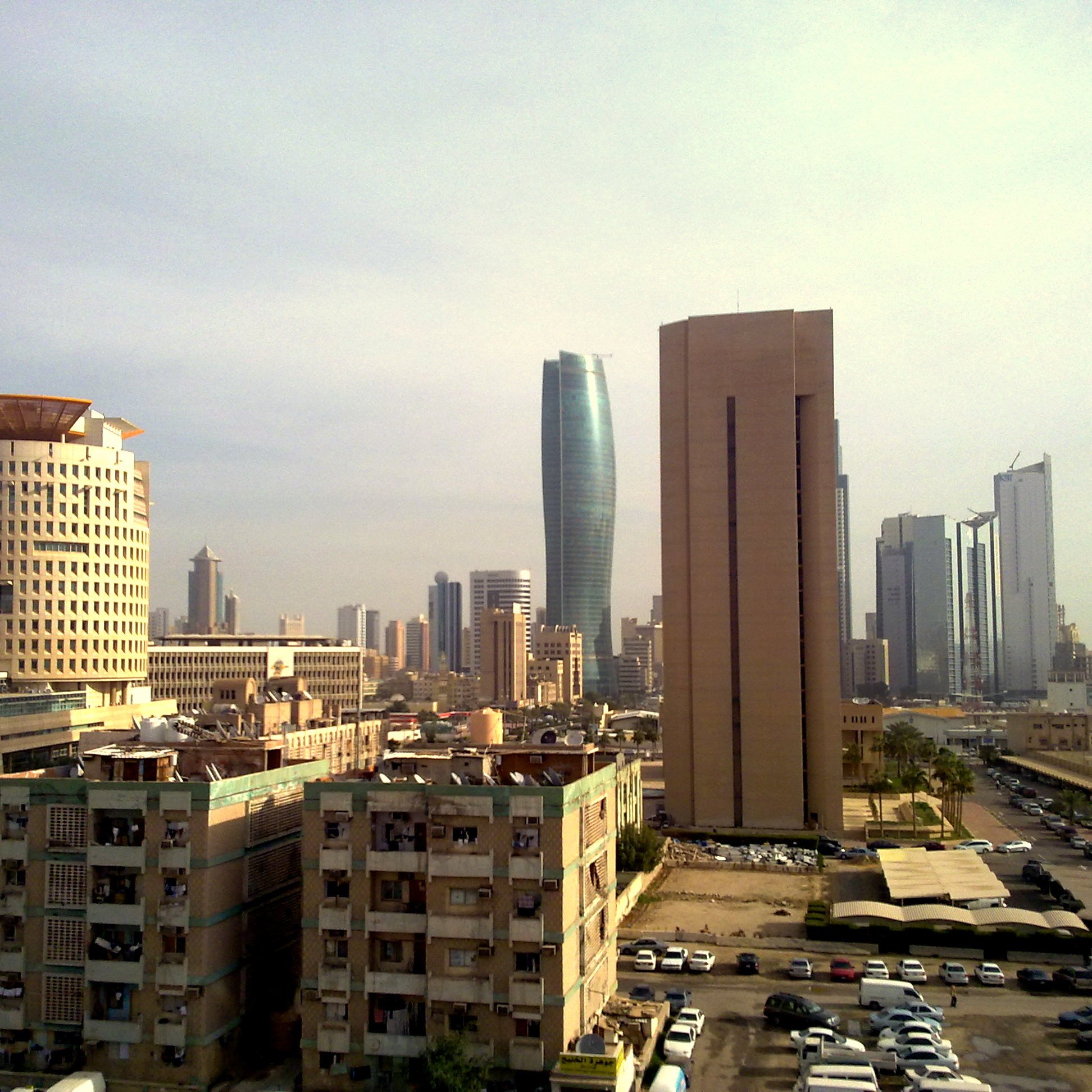 L'image montre une vue urbaine d'une grande ville moderne. On peut apercevoir plusieurs gratte-ciel aux architectures variées, avec des bâtiments à la fois anciens et contemporains. À gauche, un bâtiment au design unique, semblable à un tambour, et à droite, une structure plus élancée et moderne. En bas, on voit des immeubles vieillissants et des espaces de stationnement. Le ciel est légèrement nuageux, donnant une atmosphère calme à la scène. L'ensemble dépeint un mélange d'éléments historiques et modernes d'une métropole dynamique.