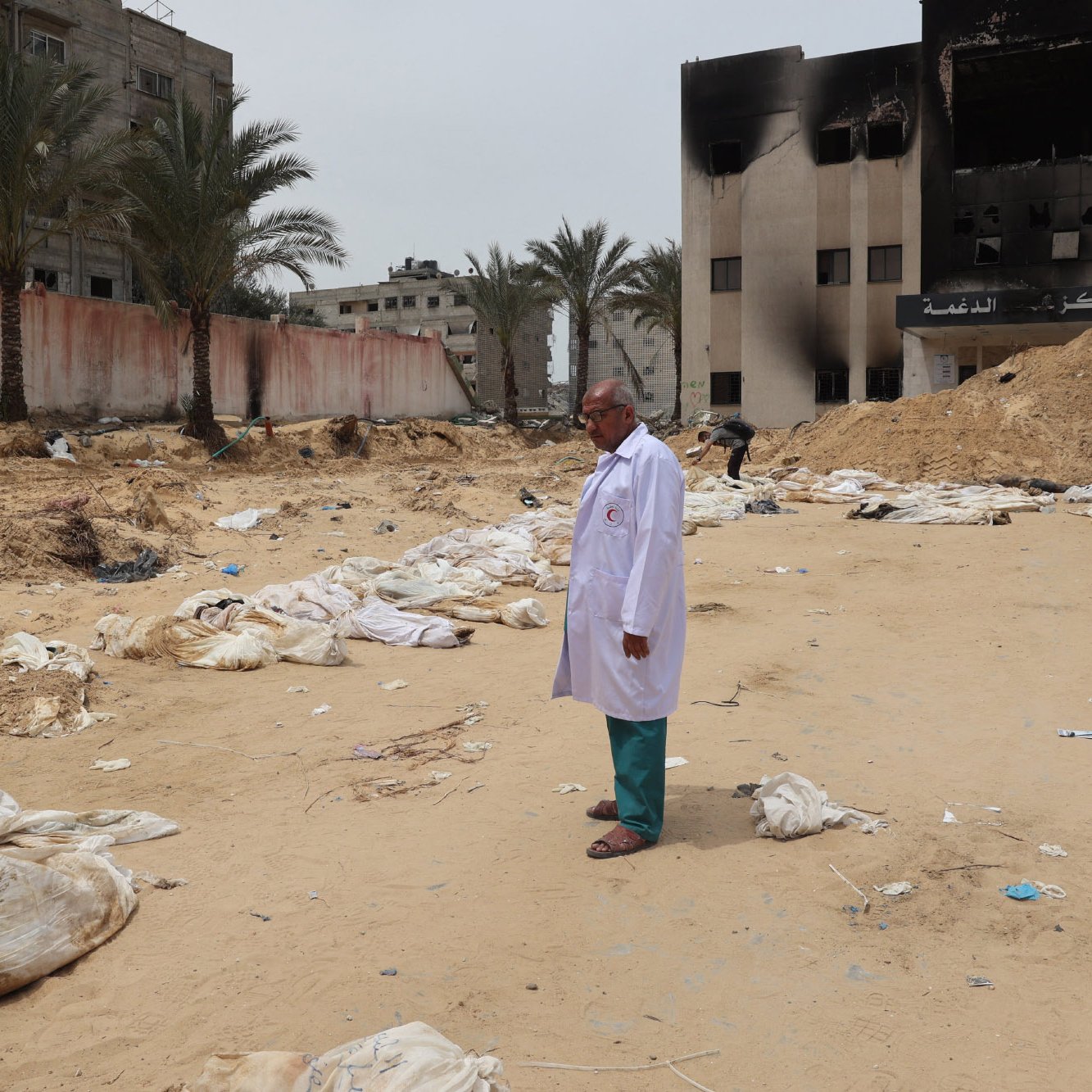L'image montre un homme en blouse blanche se tenant debout sur un terrain désertique, entouré de sacs ressemblant à des corps. En arrière-plan, on peut voir des bâtiments endommagés et des morceaux de débris éparpillés sur le sol. La scène évoque un environnement de désastre, avec une atmosphère sombre et préoccupante, suggérant des événements tragiques. Des palmiers se dressent également sur le site, ajoutant un contraste avec la gravité de la situation.