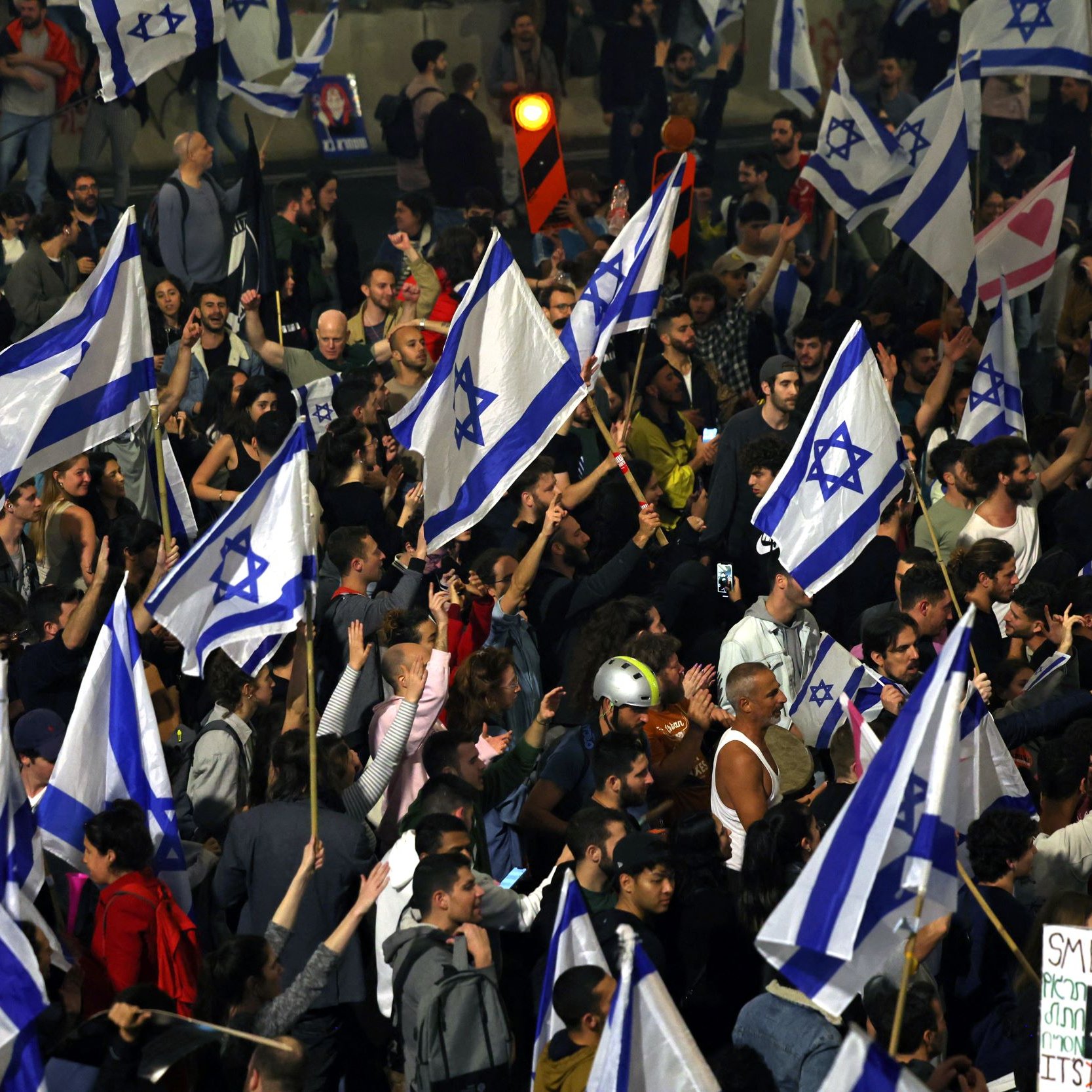 The image depicts a large crowd of people gathered together, many of whom are holding Israeli flags. The atmosphere appears vibrant and energetic, with a mix of participants visible, some raising their hands and signs. Among the signs, one reads "SMELLY C**" in a playful manner. The crowd suggests a protest or demonstration, characterized by a sense of unity and purpose, illuminated by city lights in the background.