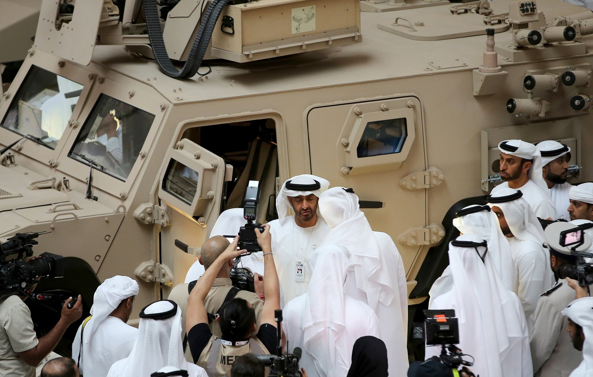 در این تصویر، گروهی از مردان به لباس سنتی عربی، در حال برگزاری یک رویداد یا نمایشگاه هستند. یکی از افراد در مرکز تصویر به نظر می‌رسد که در حال صحبت با دیگران است و چند نفر دیگر نیز در حال عکس‌برداری هستند. در پس‌زمینه، یک وسیله نقلیه نظامی با رنگ شن و طراحی خاص دیده می‌شود. این صحنه نشان‌دهنده یک موقعیت رسمی و احتمالا نظامی است، با حضور رسانه‌ها و توجه به جزئیات.