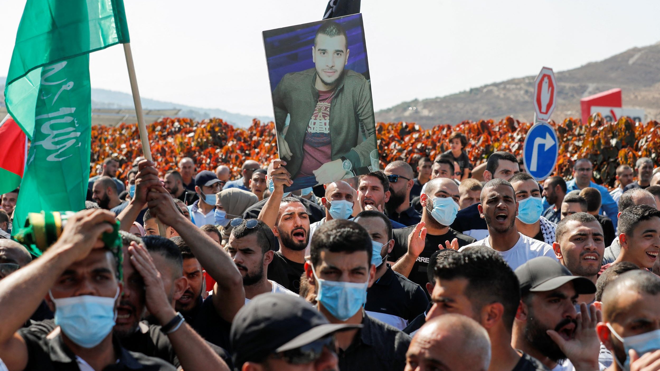 La imagen muestra a un grupo de personas en una manifestación. Hay banderas de diferentes colores ondeando, y muchos de los manifestantes llevan mascarillas. Uno de ellos sostiene una fotografía en alto de un hombre. El ambiente parece ser de protesta, y el fondo incluye áreas despejadas con vegetación. La escena transmite una fuerte sensación de unidad y determinación entre los participantes.