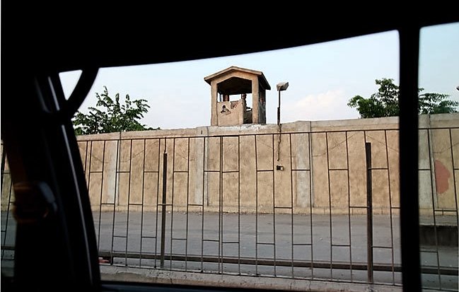 L'image montre un mur de béton surmonté d'une petite structure, probablement un poste de garde, avec un toit en pente. Au premier plan, on peut voir une grille métallique qui sépare l'intérieur de la scène extérieure. En arrière-plan, des arbres verdoyants apparaissent, ajoutant une touche de nature à l'environnement urbain. La lumière de la fin de journée crée une atmosphère calme.