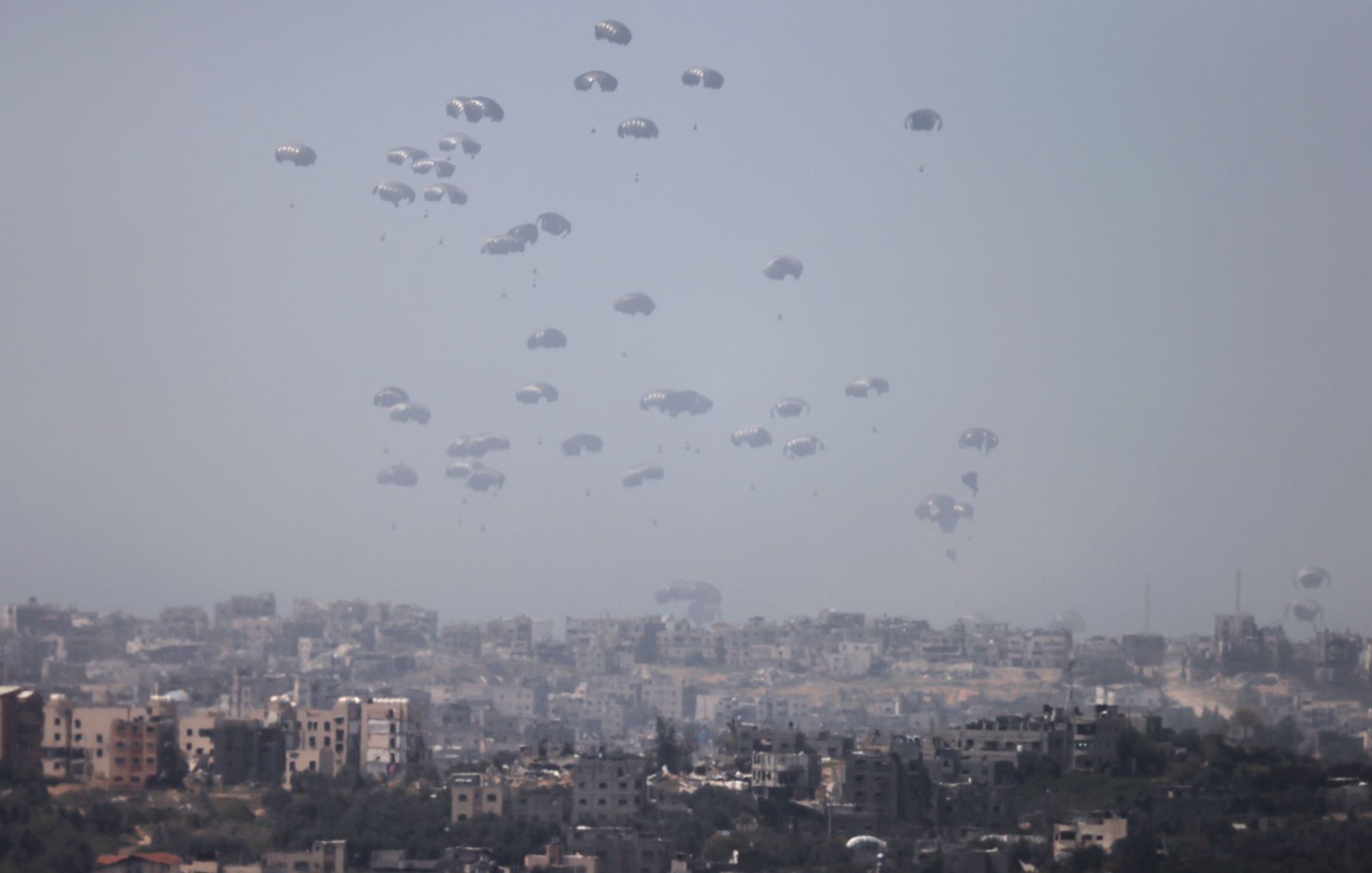 در این تصویر، چندین چتر نجات در آسمان به طور هم‌زمان دیده می‌شوند که به آرامی در حال سقوط هستند. زمینه تصویر شهری است که شامل ساختمان‌ها و محله‌های مختلف است. آسمان ابری و کمی تاریک به نظر می‌رسد و این باعث می‌شود فضای تصویر حالتی جدی و نگران‌کننده داشته باشد. تصویر به خوبی نمایانگر یک وضعیت نظامی یا اضطراری است.