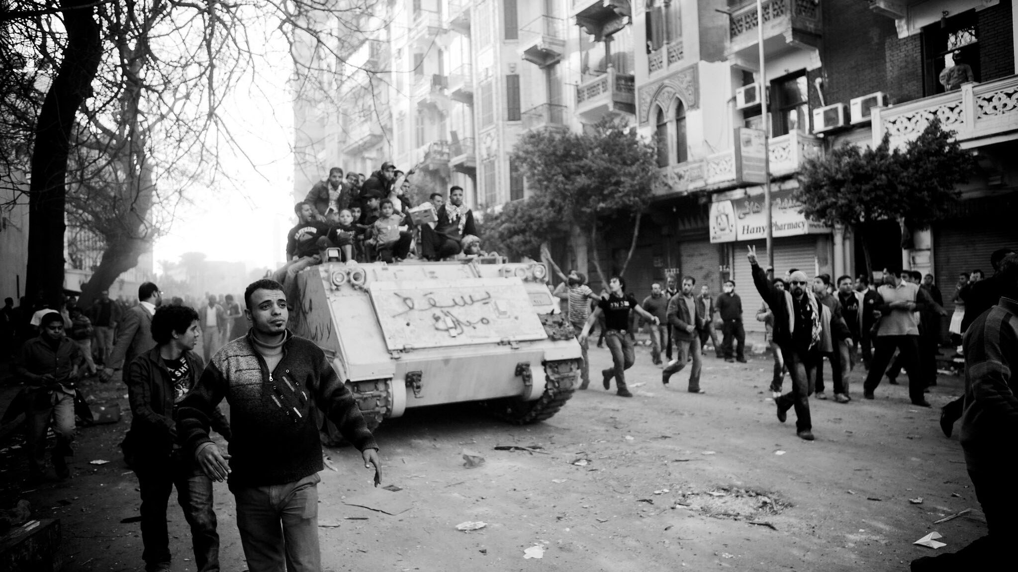 L'image montre une scène de manifestation urbaine, avec des gens dans une rue animée. Au premier plan, plusieurs manifestants marchent, certains levant les bras en l'air. À l'arrière-plan, un tank est visible, portant des inscriptions. La scène est en noir et blanc, ce qui intensifie l'atmosphère dramatique et chargée d'émotion. Les bâtiments environnants et les arbres ajoutent à l'environnement urbain. L'ensemble dégage un sentiment de tension et de mouvement.