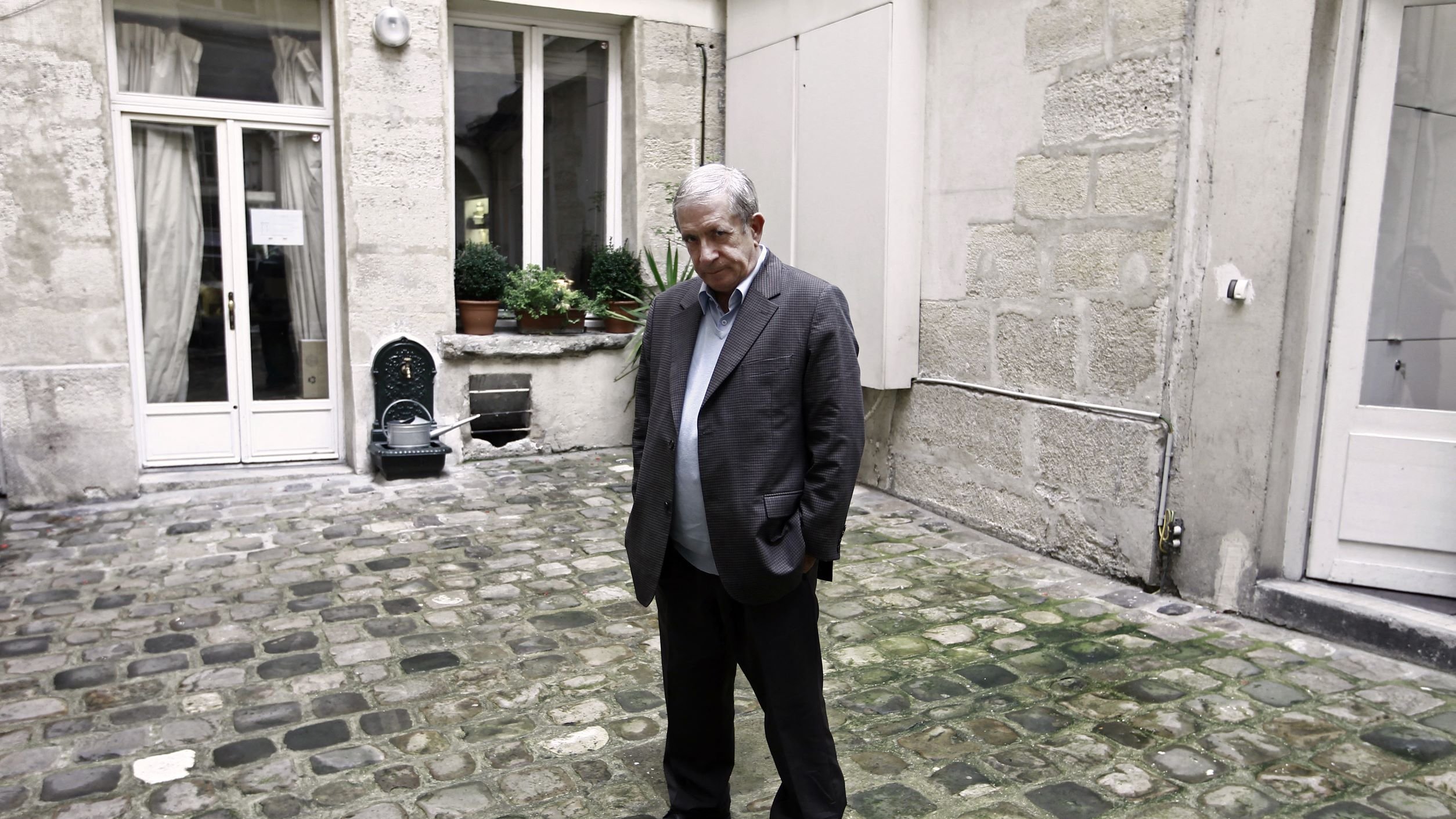 L'image montre un homme debout dans une cour pavée, visiblement pensif ou mélancolique. Il porte un costume brun et se tient les mains dans les poches. L'arrière-plan est constitué de murs en pierre et de fenêtres, avec quelques plantes en pot. L'atmosphère semble calme, mais un peu triste.