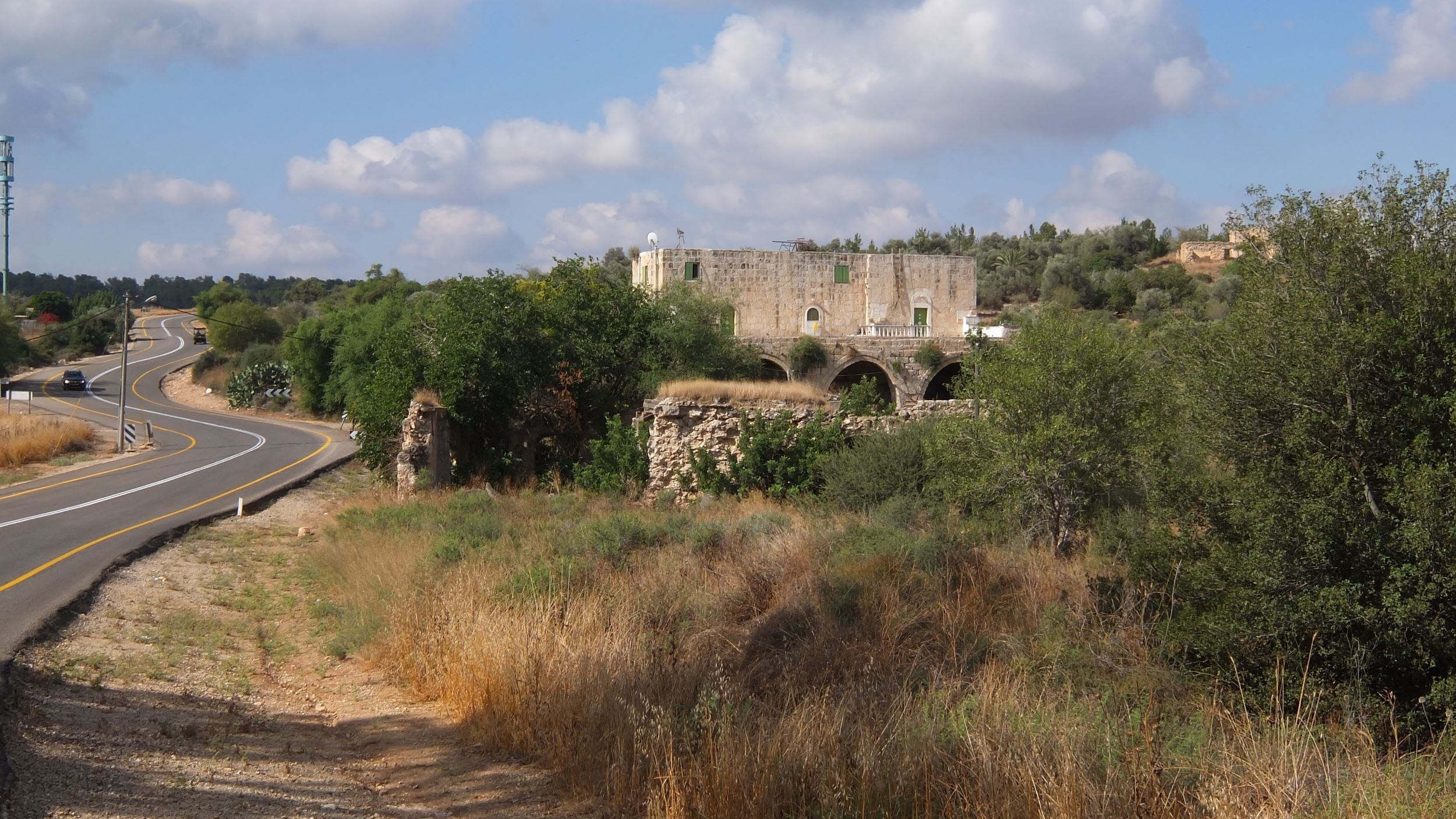 L'immagine mostra un paesaggio rurale caratterizzato da una strada curvilinea che si snoda tra la vegetazione. Ai lati della strada, ci sono prati e aree erbose, con un'atmosfera tranquilla e naturale. Sullo sfondo, spicca una vecchia costruzione in pietra, apparentemente in rovina, con archi visibili, circondata da alberi e vegetazione. Nel cielo, si possono vedere alcune nuvole sparse, che completano il quadro di un ambiente sereno e suggestivo.