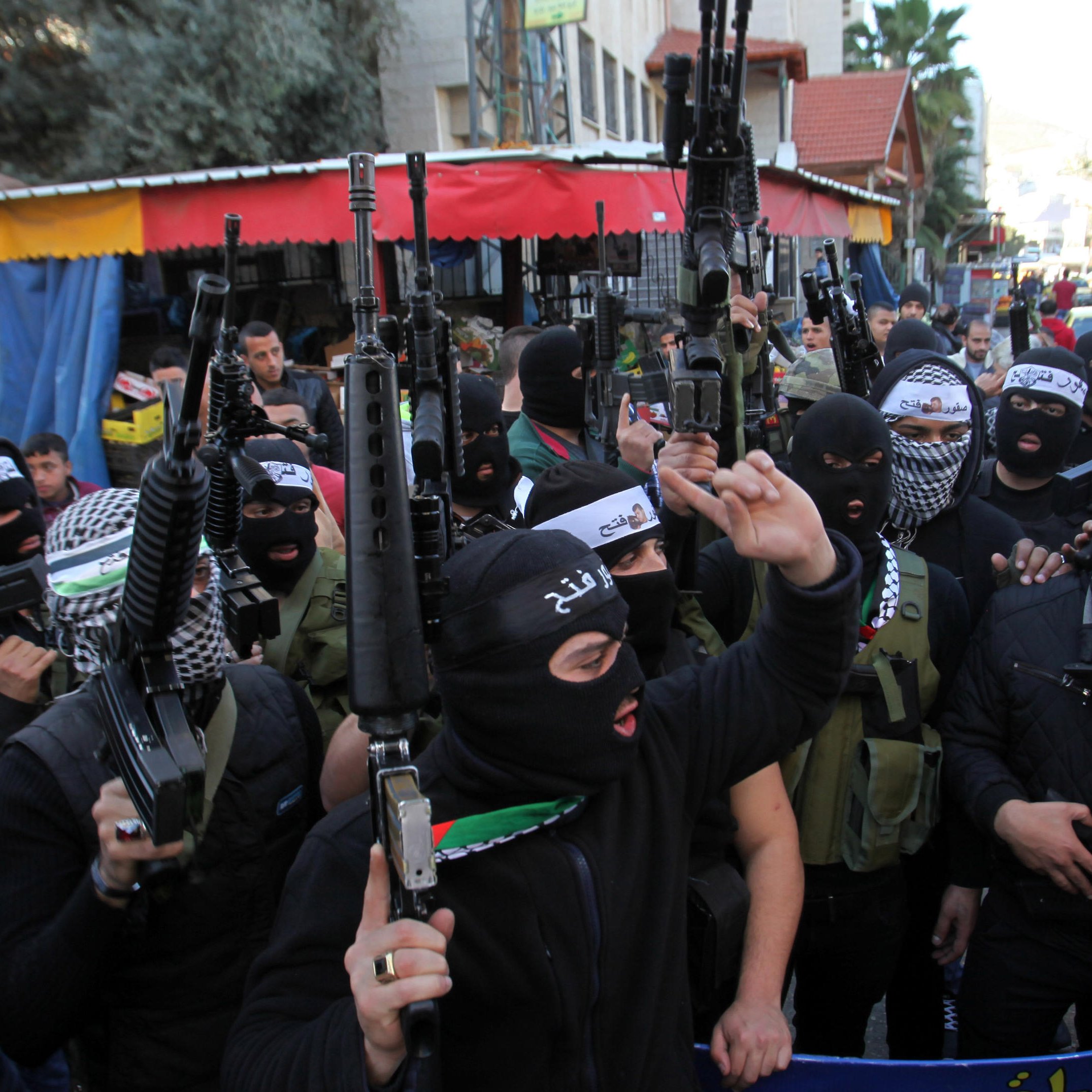 L'image montre un groupe de personnes portant des masques noirs et tenant des armes. Elles se rassemblent dans une rue, avec des bâtiments et des commerçants en arrière-plan. L'atmosphère semble tendue, et leurs expressions indiquent une certaine détermination ou colère. Des drapeaux et des banderoles sont visibles, suggérant un contexte de manifestation ou de rassemblement politique.