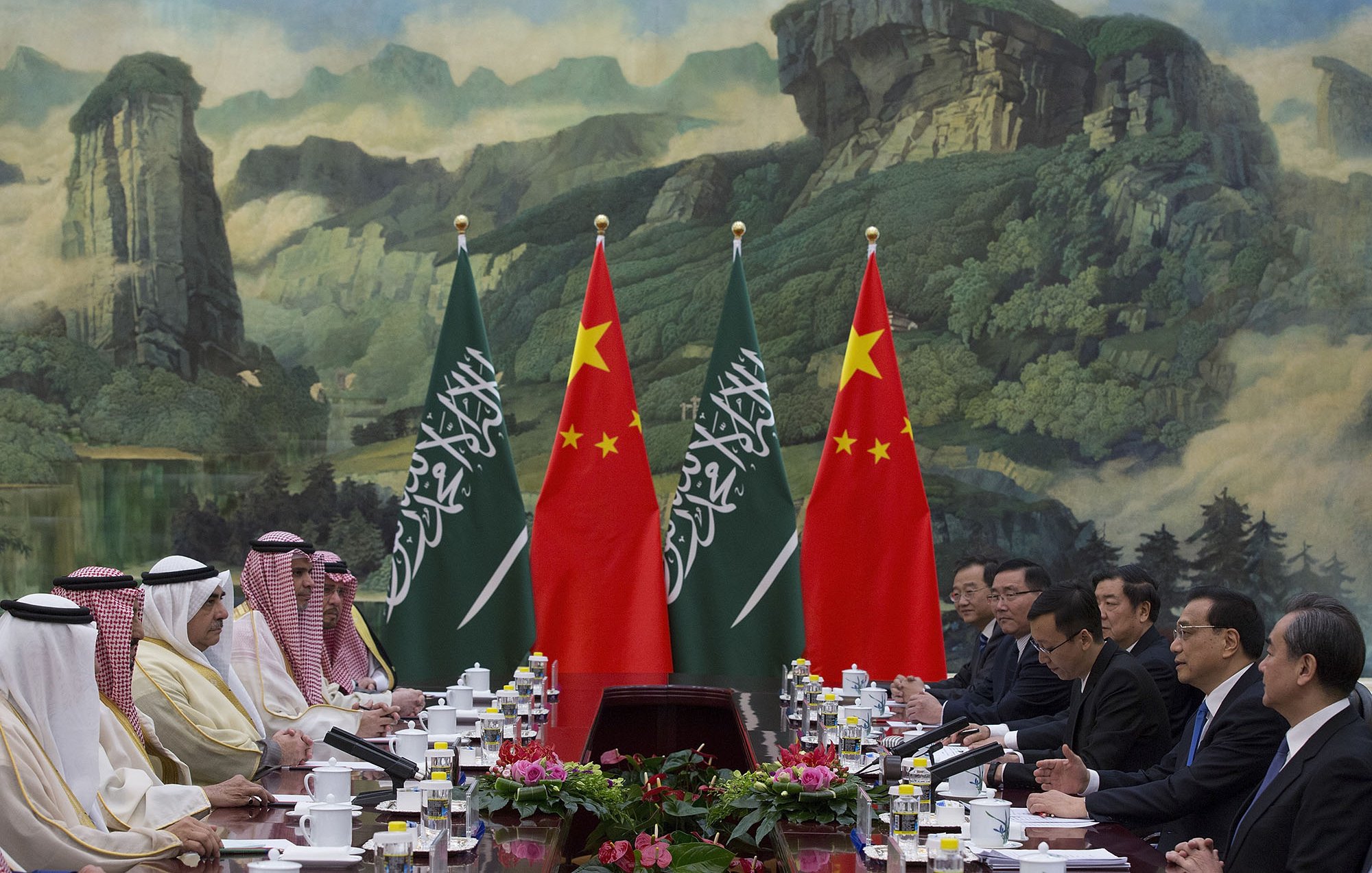 L'image montre une réunion diplomatique entre des représentants de l'Arabie saoudite et de la Chine. Les participants sont assis à une table, entourés de drapeaux des deux pays. En arrière-plan, on peut voir une grande fresque représentant des paysages naturels. La scène dégage une ambiance formelle, typique des sommets ou des négociations internationales. Sur la table, il y a des documents et des tasses de thé.