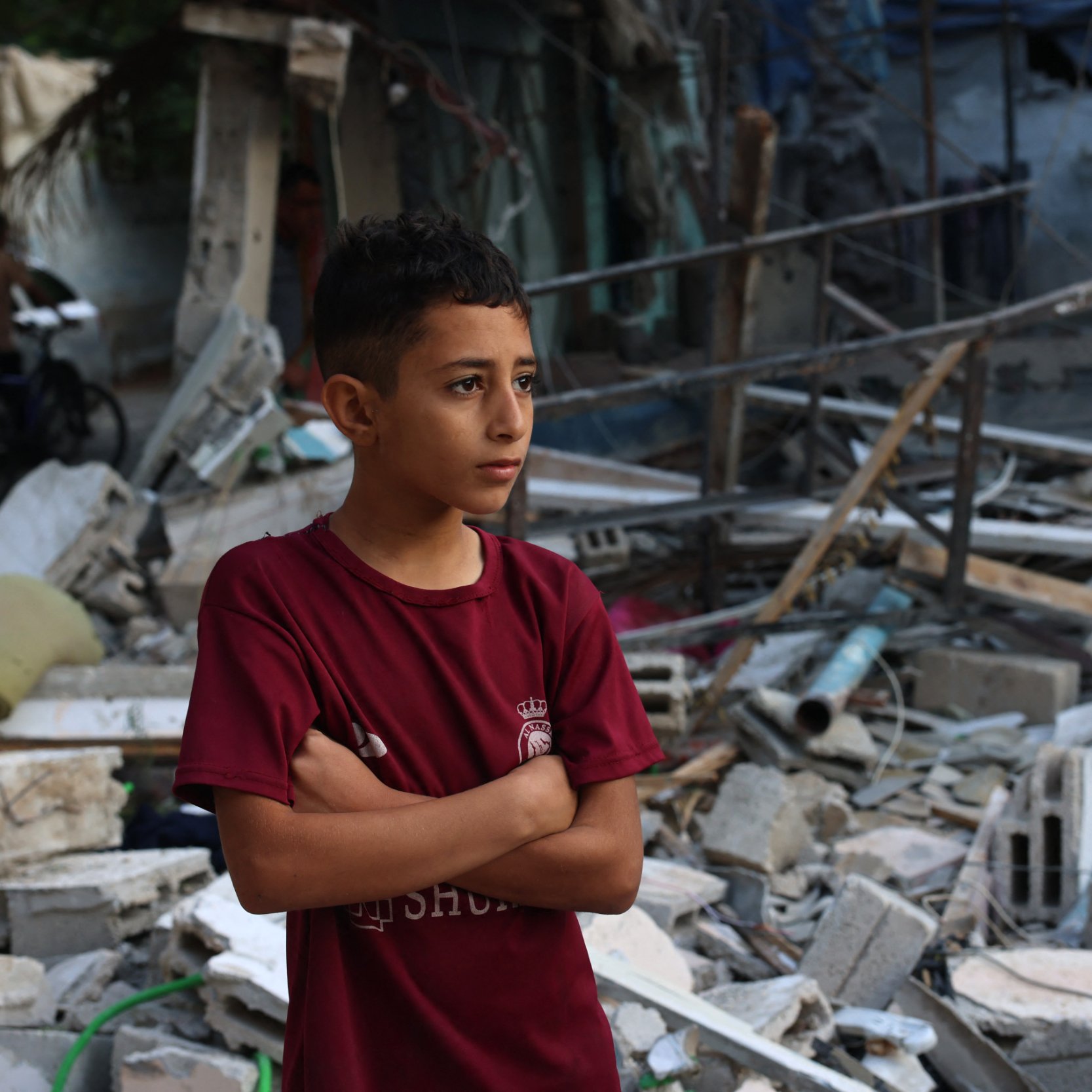 L'immagine mostra un ragazzo con le braccia incrociate, che sembra riflettere o osservare attentamente. Sullo sfondo, ci sono macerie e detriti, suggerendo che si trova in un'area devastata, probabile frutto di un conflitto o di una calamità. Il suo sguardo serio e pensieroso comunica un senso di tristezza e resilienza di fronte a una situazione difficile. La scena evidenzia anche la vulnerabilità dei bambini in contesti di crisi.