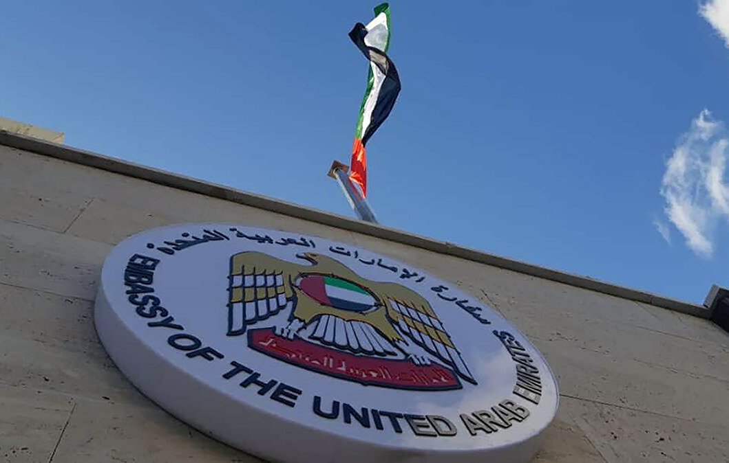 L'image montre un bâtiment avec un panneau qui indique qu'il s'agit de l'ambassade des Émirats arabes unis. Au-dessus du panneau, on peut voir le drapeau des Émirats arabes unis flottant. Le ciel est clair et légèrement nuageux, ce qui donne une belle arrière-plan à la scène.