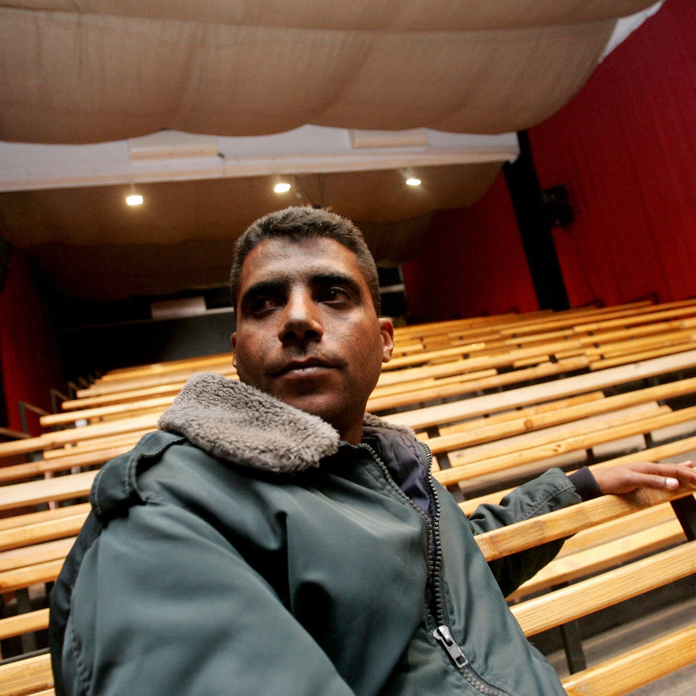 L'immagine mostra un giovane uomo seduto su una panca di legno in un teatro. Il teatro ha un'illuminazione soffusa, con tende rosse che coprono le pareti e un palcoscenico visibile sullo sfondo. L'uomo indossa una giacca scura con un colletto di pelliccia. La sua espressione sembra seria e contemplativa mentre guarda verso l'esterno, evidenziando un'atmosfera di introspezione nel contesto di un luogo culturale. Le file di sedili vuoti suggeriscono un ambiente che sta aspettando un evento o una performance.