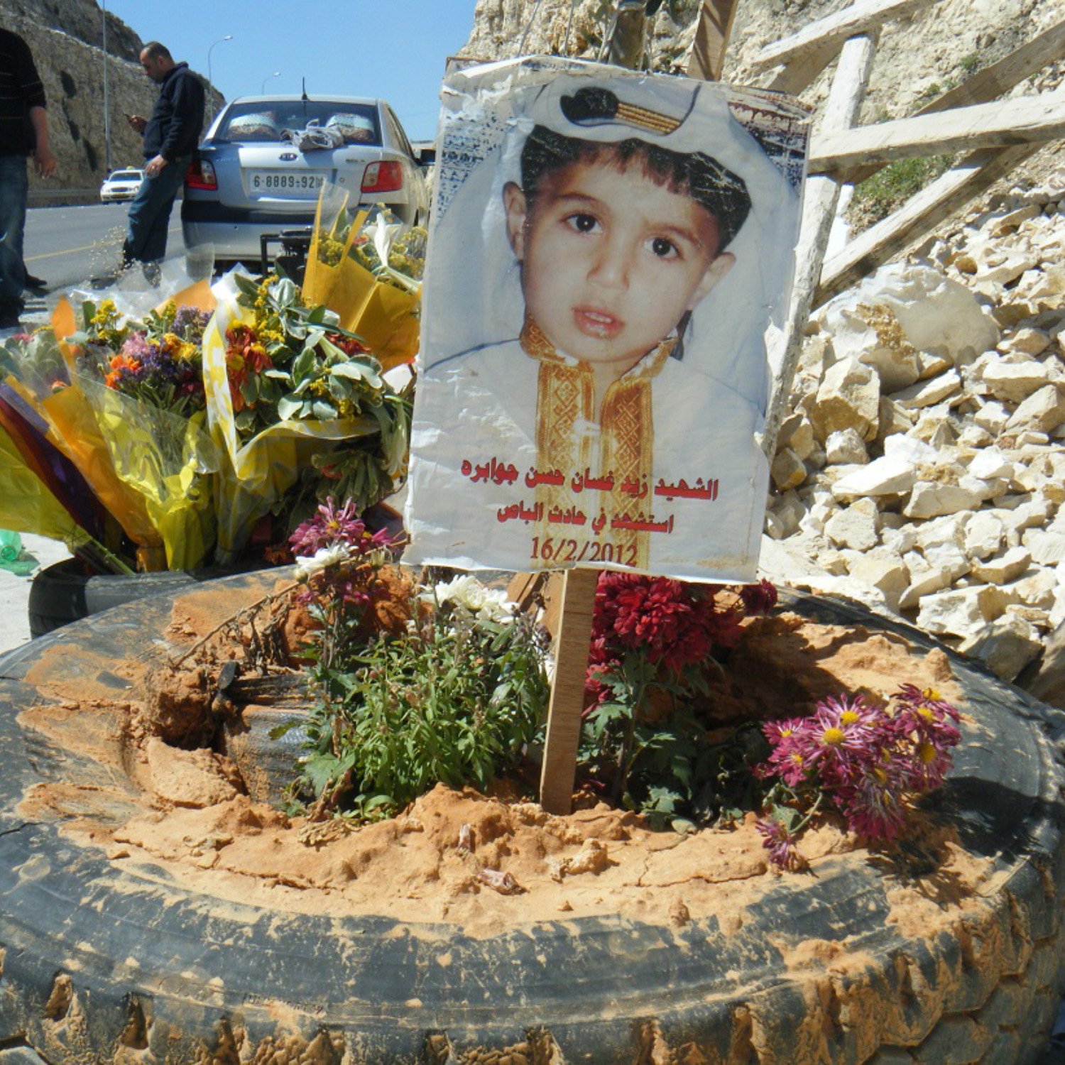 L'image montre un mémorial pour un enfant, avec une photo de lui affichée sur un panneau en bois planté dans un pneu. Autour du panneau, il y a des fleurs multicolores et de la terre. En arrière-plan, on peut voir des personnes rassemblées et un paysage de pierre. Cela semble être un hommage émouvant à un enfant décédé.