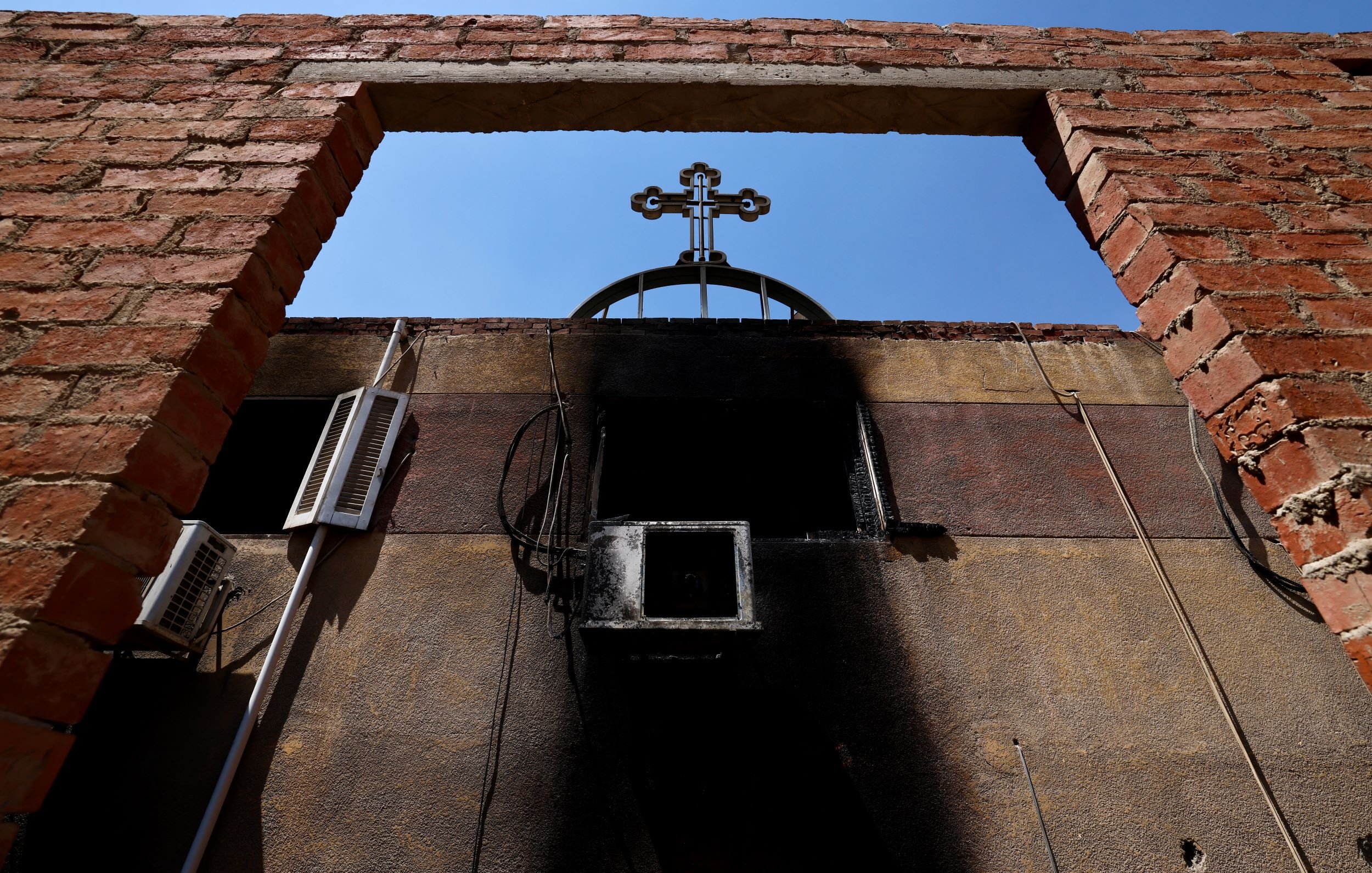 L'image montre un cadre en briques laissant entrevoir un ciel bleu, avec une croix noire au sommet d'une structure. On peut également voir des éléments d'électricité sur le mur, et une partie du mur semble avoir subi des dommages dus à un incendie, avec des marques noires. L'ensemble dégage une atmosphère de contraste entre la lumière et l'ombre, ainsi qu'une impression de désolation.